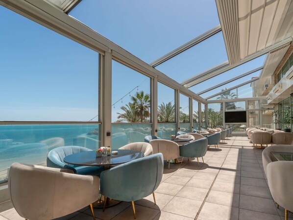 Un espace extérieur lumineux, aménagé pour accueillir les clients, avec vue sur l’océan, comprenant des tables rondes et des chaises modernes.