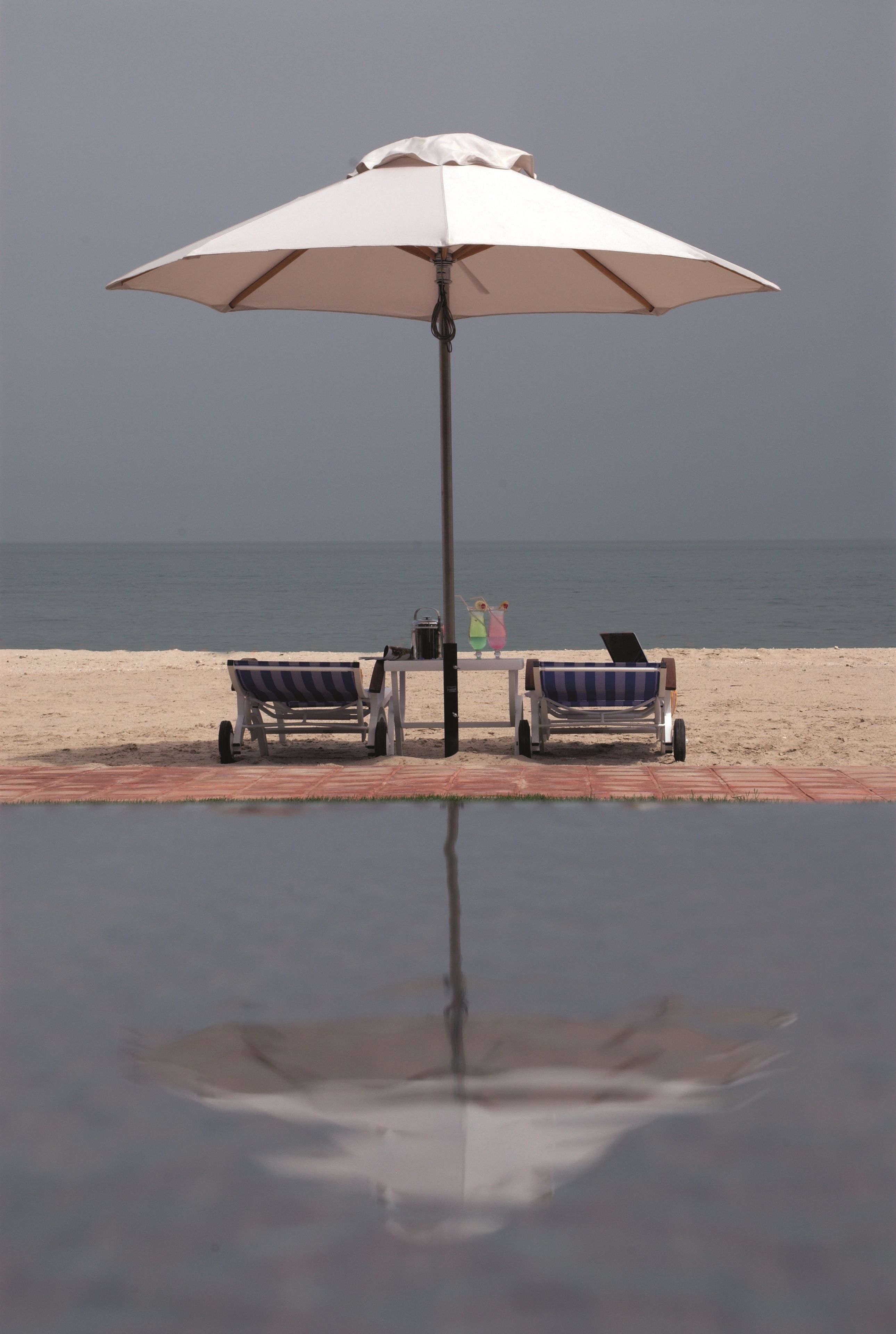 Zwembad aan het strand met twee ligbedden en een tafel onder een grote parasol.