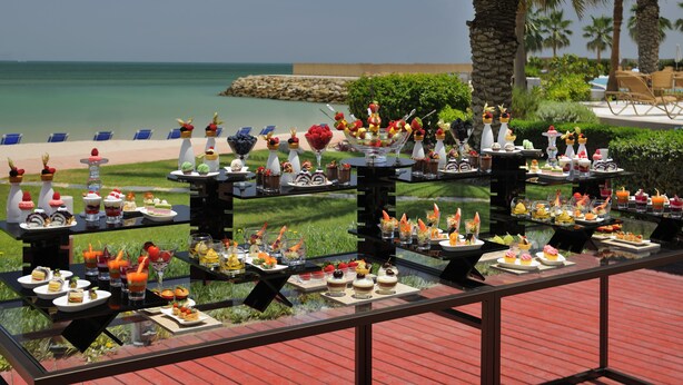 Une table de buffet proposant divers plats et boissons, le tout installé sur fond de plage et de palmiers.