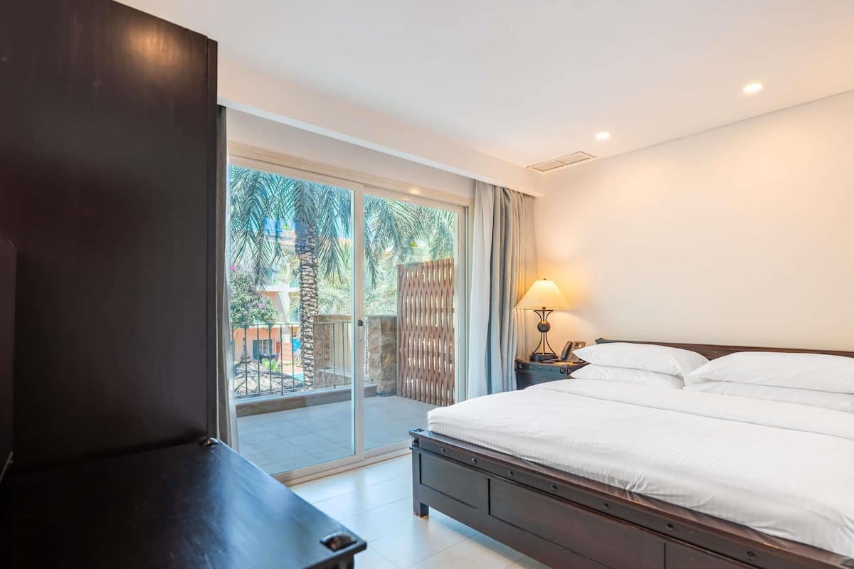 Ein Hotelzimmer mit Bett, Fernseher, Lampe und Blick auf einen Innenhof mit Palmen.