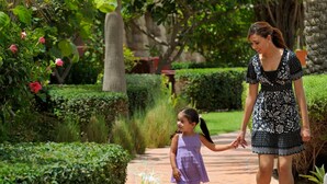 Une femme et un enfant se promènent sur un chemin pavé, dans un jardin luxuriant agrémenté de palmiers et de haies bien entretenues.