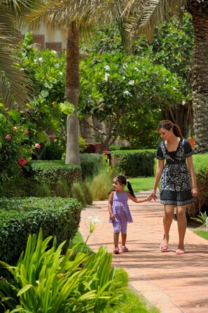 Una mujer y un niño caminando por un camino pavimentado en un jardín exuberante con palmeras y setos cuidados.