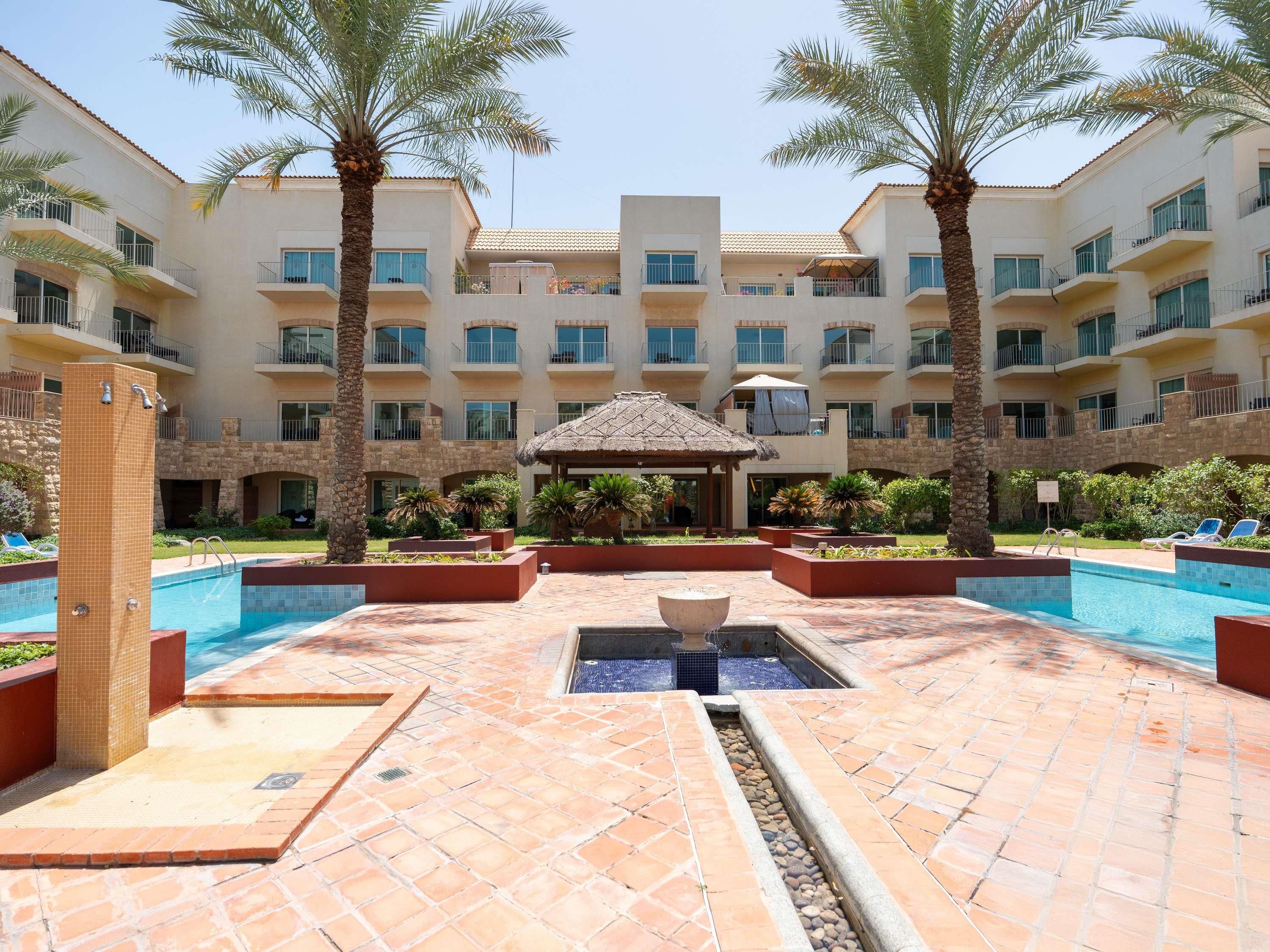 Ein Hotelinnenhof mit Swimmingpool, Palmen und einem Gazebo mit Reetdach.