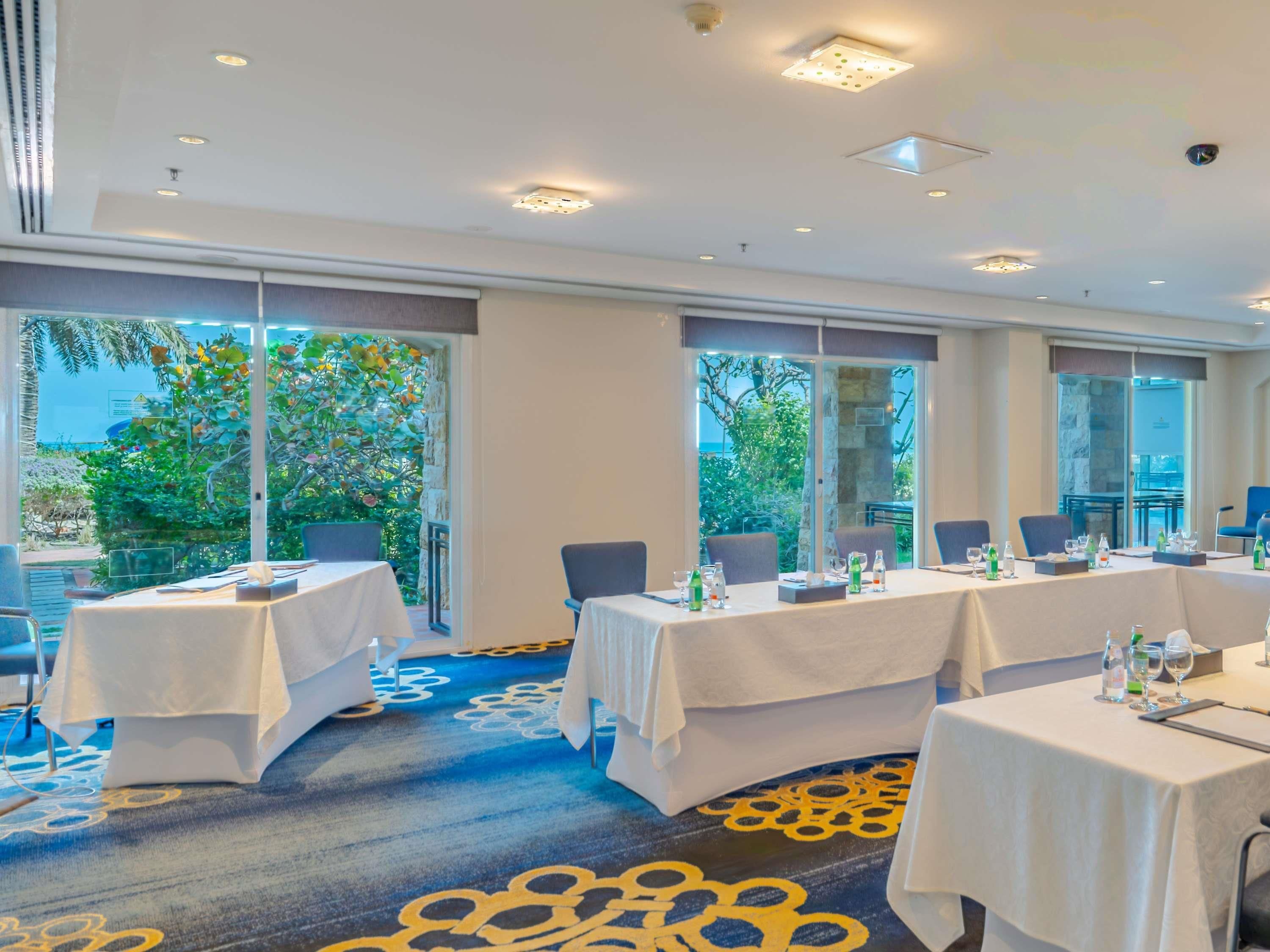 Un salón de conferencias con mesas, sillas y vistas al exterior con vegetación.