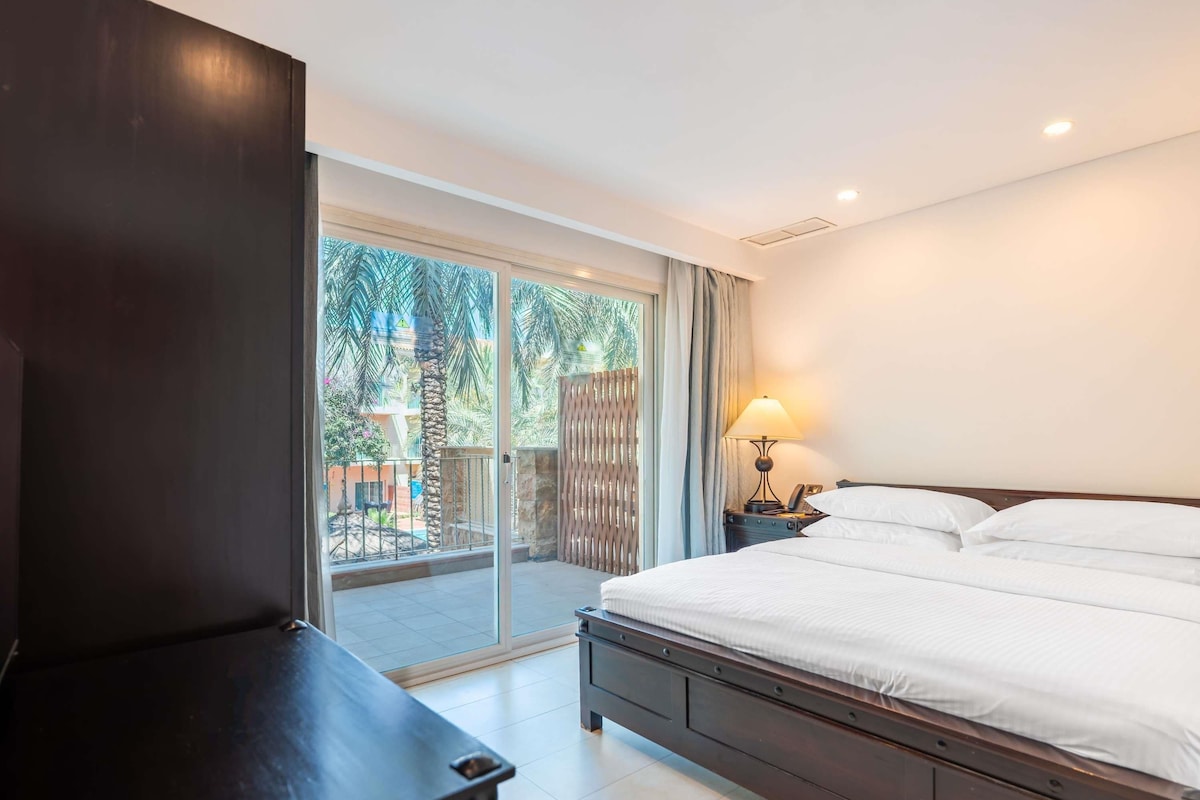 Una habitación de hotel con cama, televisión, lámpara y vista a un patio con palmeras.