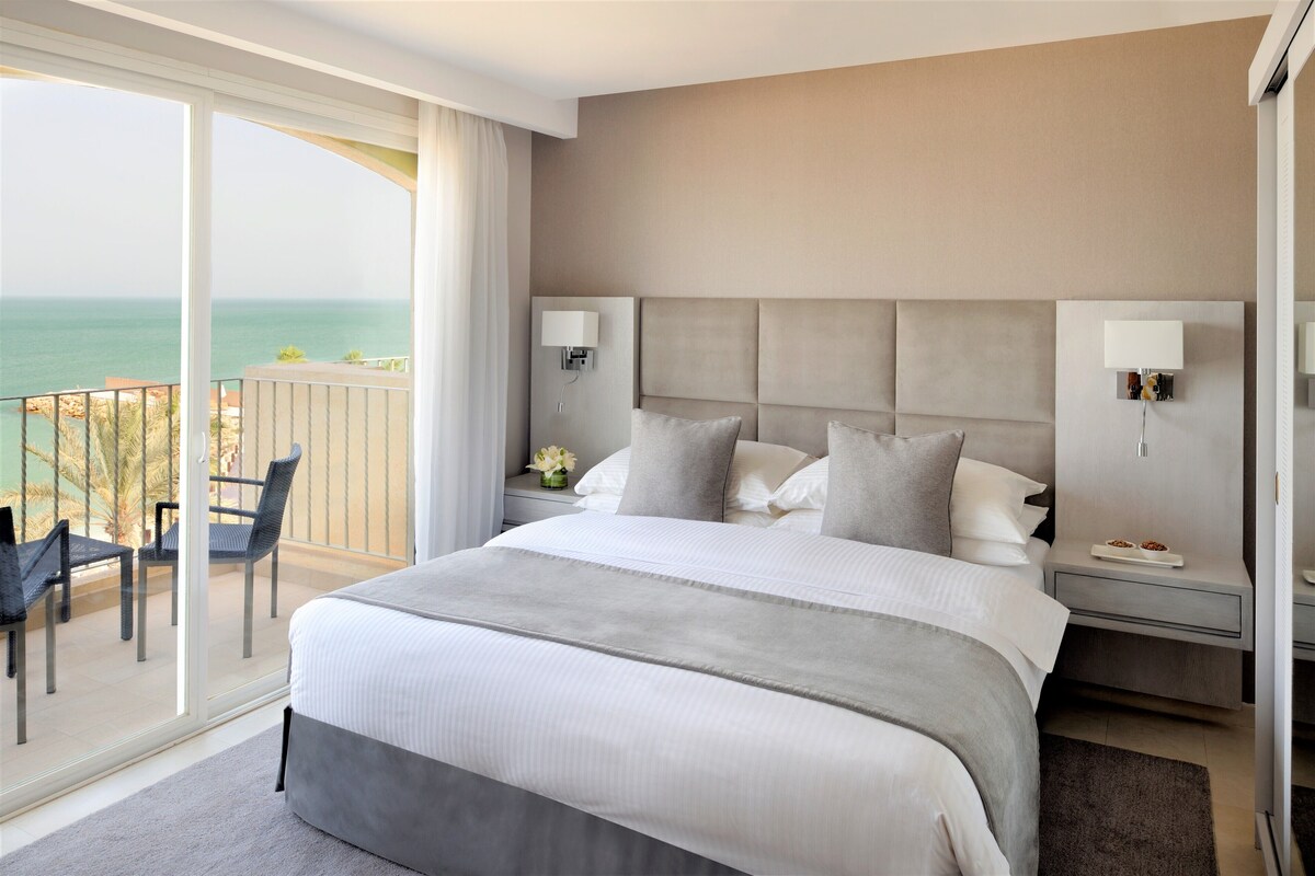 Una habitación de hotel con una cama grande, dos mesitas de noche, un balcón con sillas y vista al mar.