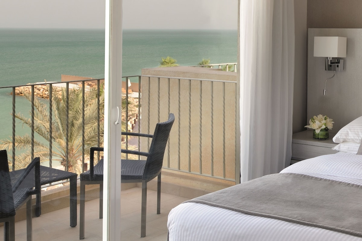 Habitación de hotel con balcón que da al mar, una cama, una mesita de noche con lámpara y una silla.