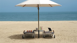 Escena en la playa con una sombrilla blanca, dos sillas de descanso, una mesa con bebidas y un ordenador portátil.