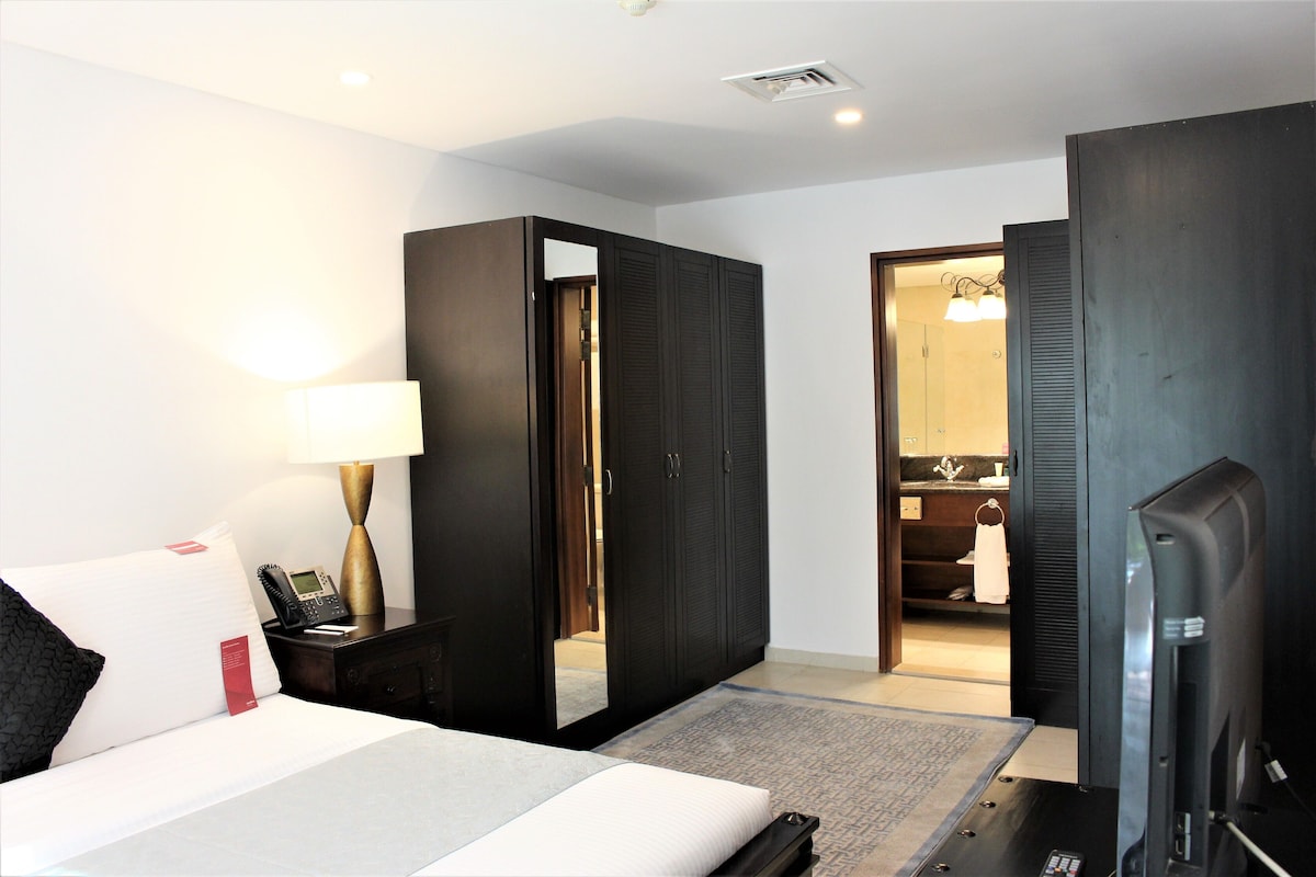 Una habitación de hotel con cama, lámpara de noche, televisor y espejo.