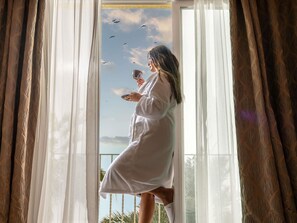 A person in a white robe standing by a window with a view of birds and a clear sky.