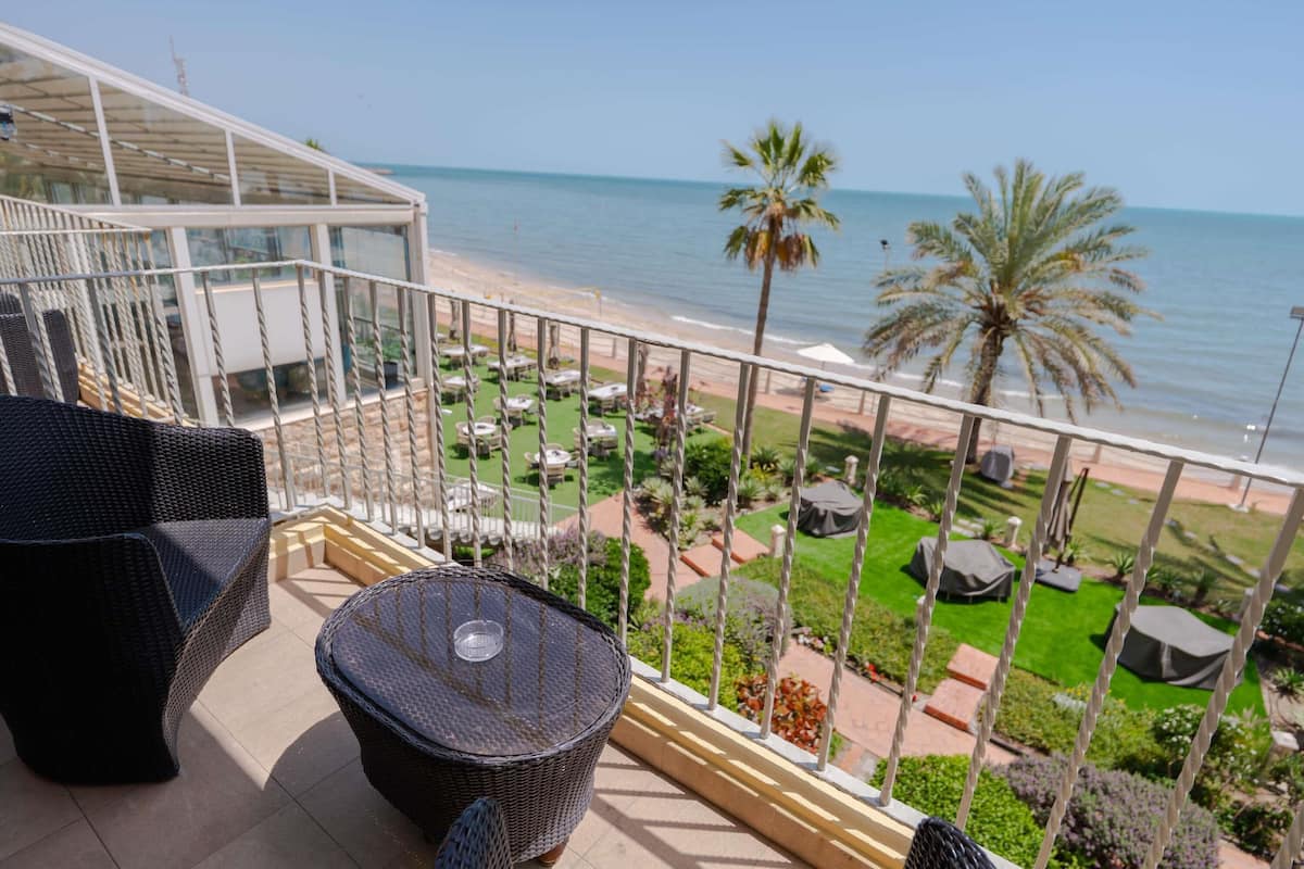 Ein Balkon mit Korbmöbeln, einer Glaskonstruktion und Blick auf den Strand und Palmen.