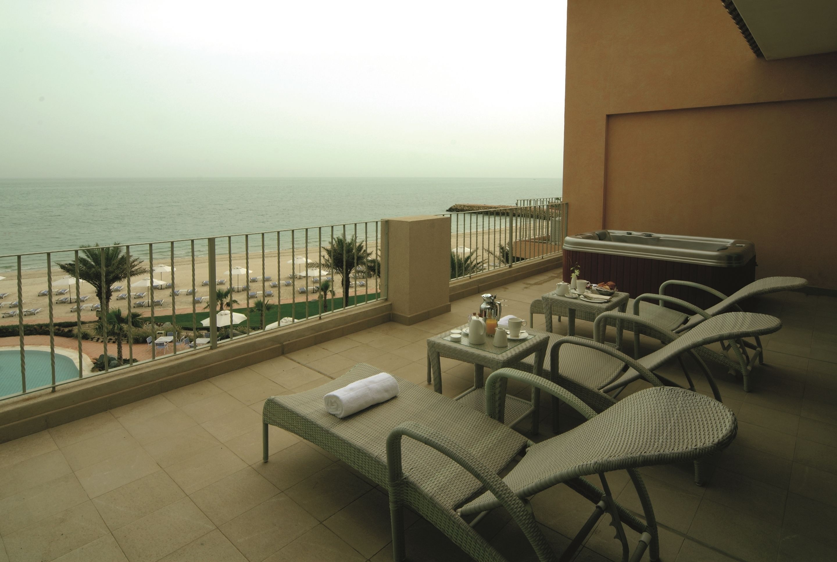 A balcony with a view of the beach, lounge chairs, a table with a kettle, and a small pool.