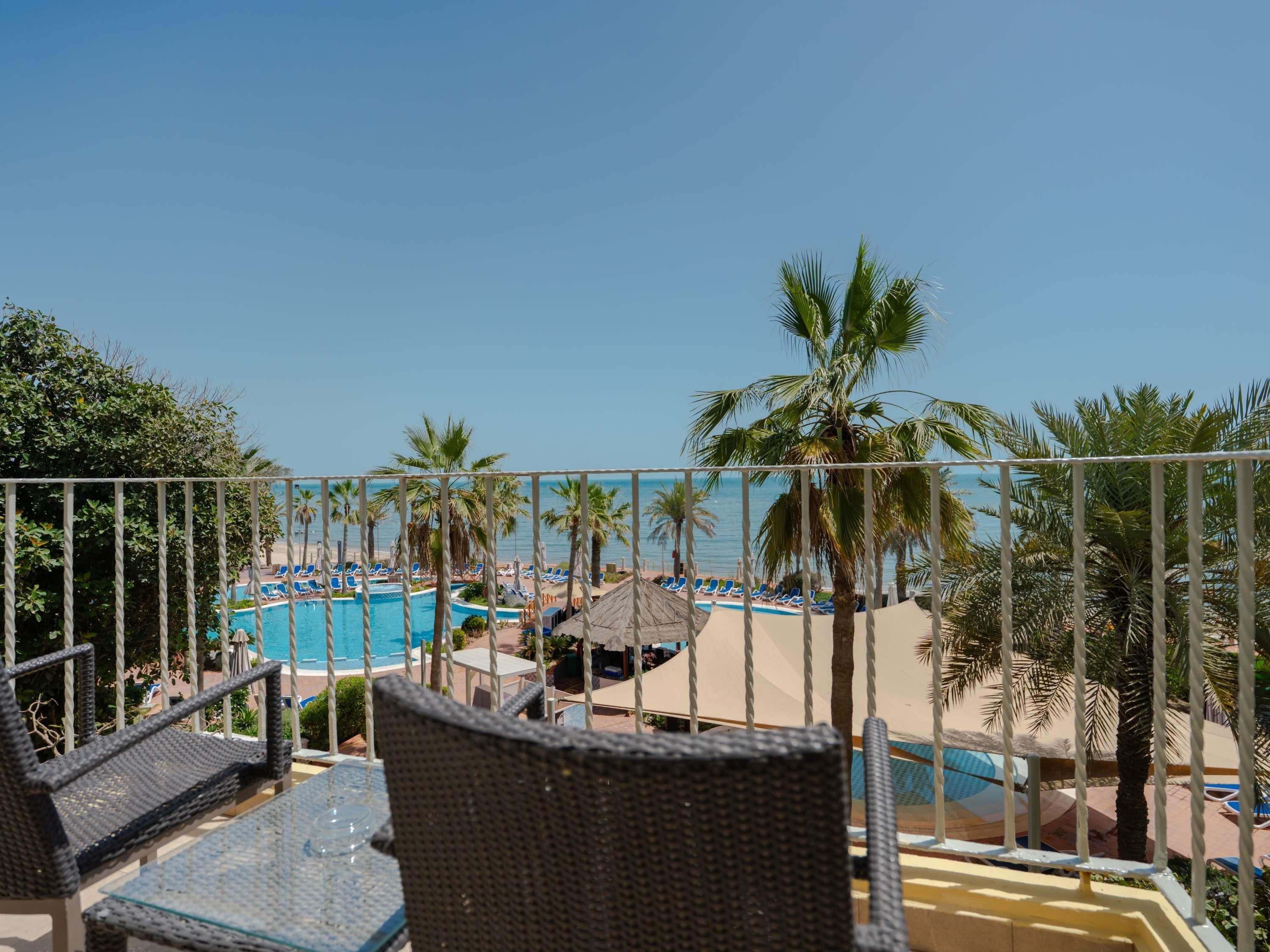 A balcony with wicker chairs and a glass table overlooking a pool area with lounge chairs and palm trees.