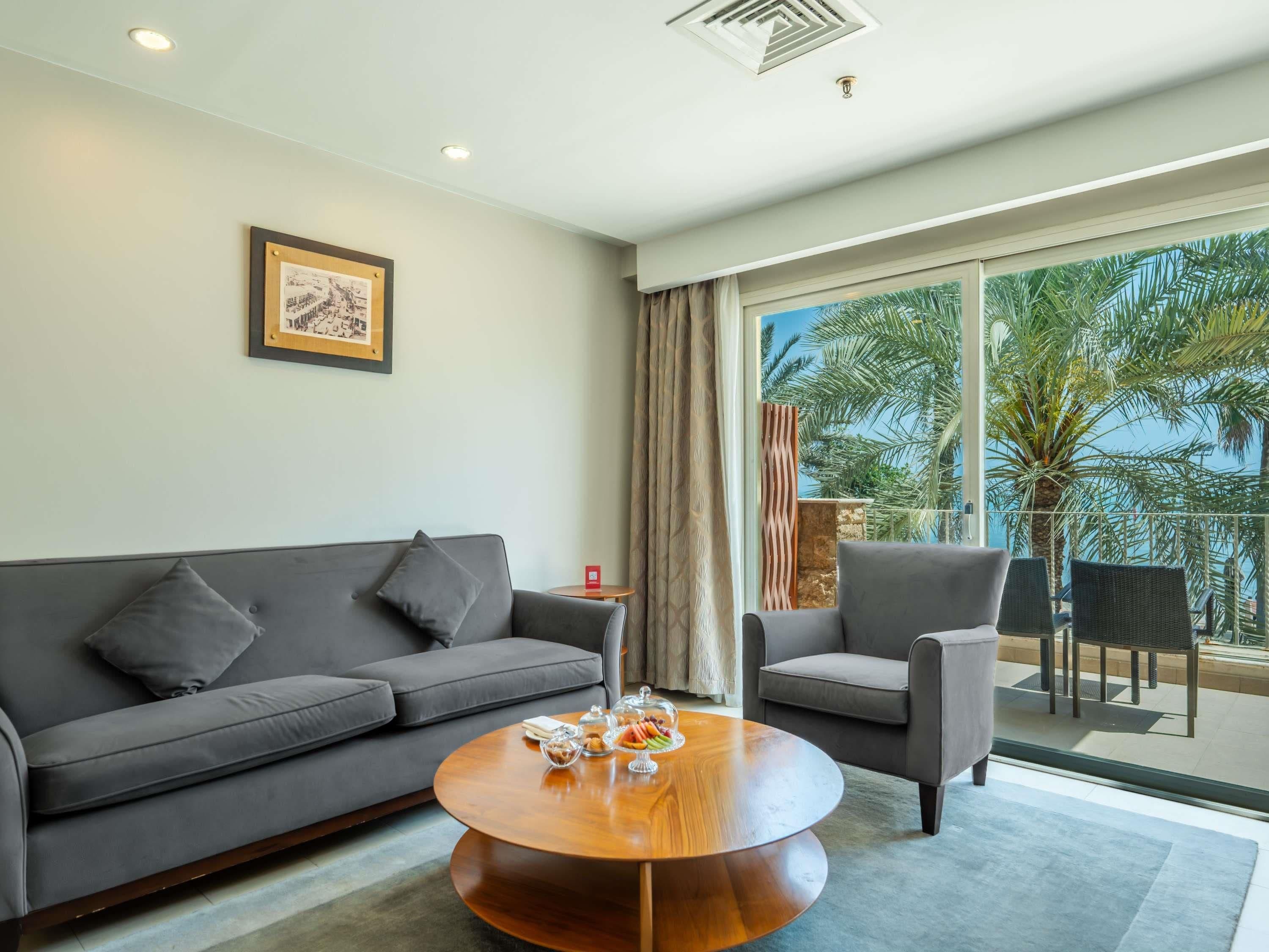 Un soggiorno moderno con un divano grigio, un tavolino rotondo con oggetti decorativi e vista su palme attraverso ampie porte scorrevoli in vetro.