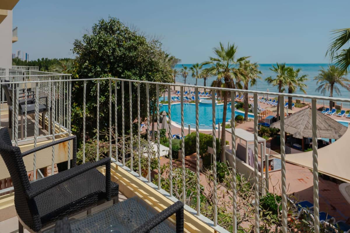 Ein Balkon mit Blick auf Pool und Strand, umgeben von Palmen und Liegestühlen.