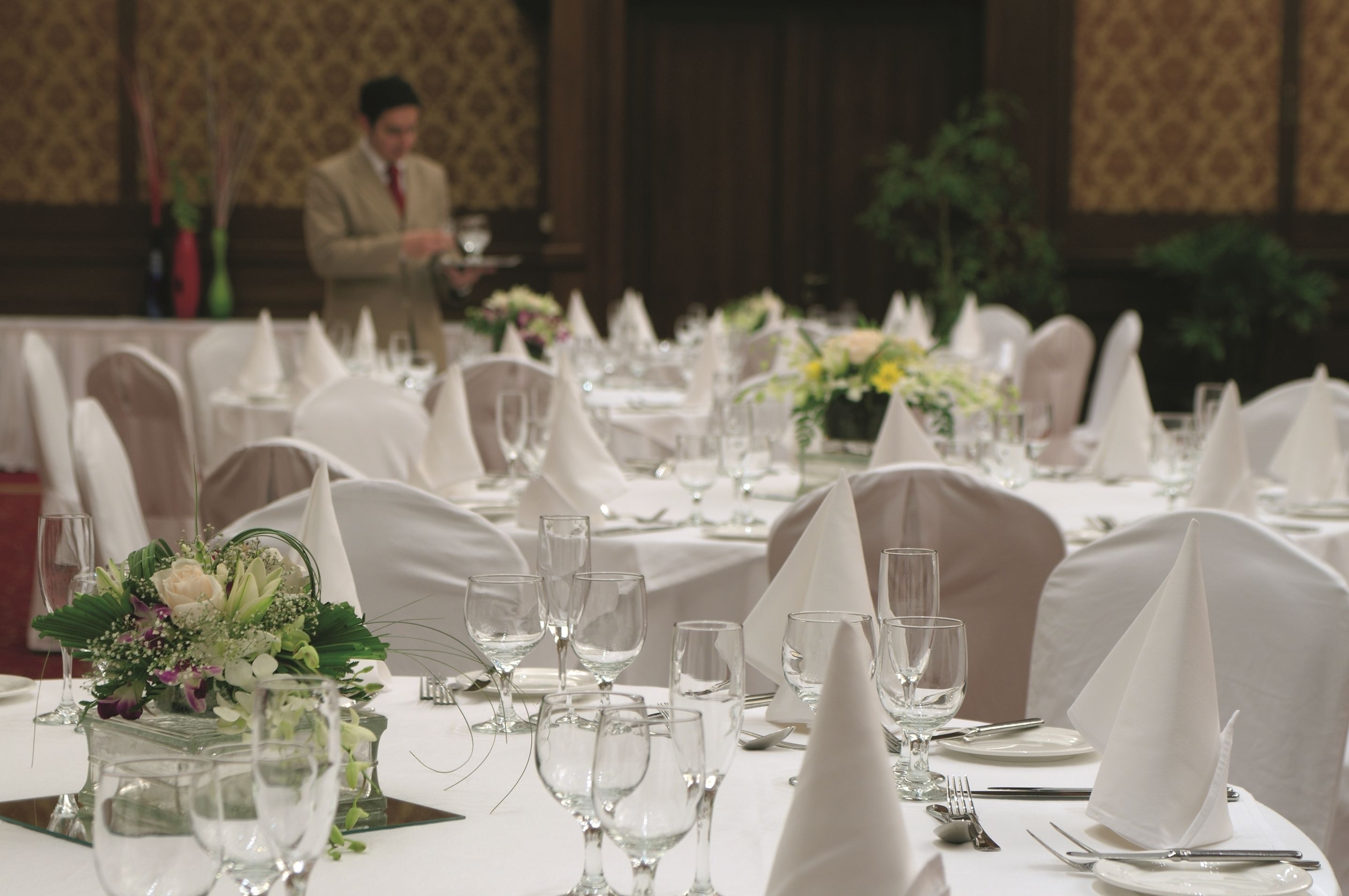 A banquet hall with round tables set for a formal event, each with white tablecloths, glassware, and floral centerpieces.
