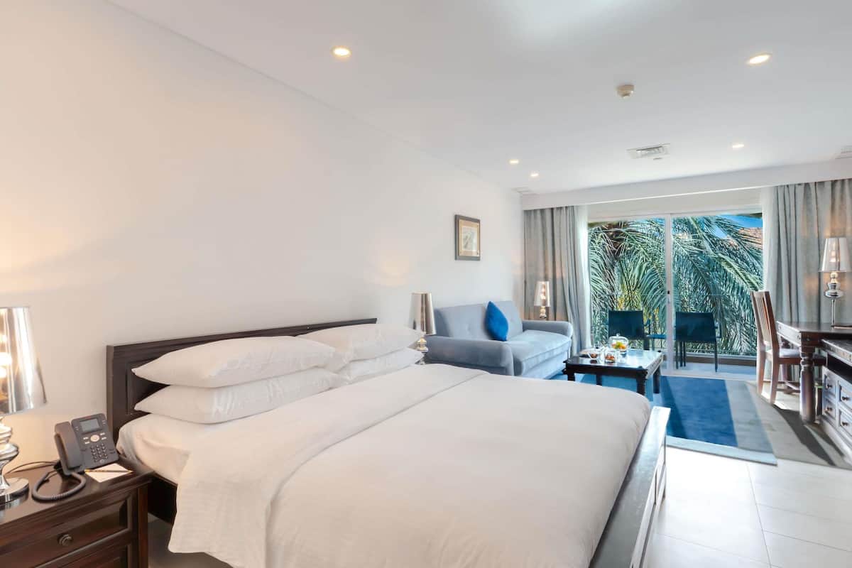 Ein Hotelzimmer mit einem großen Bett, Nachttischtelefon, einem Schreibtisch mit Lampe, einem Sessel und Blick auf Palmen durch das Fenster.