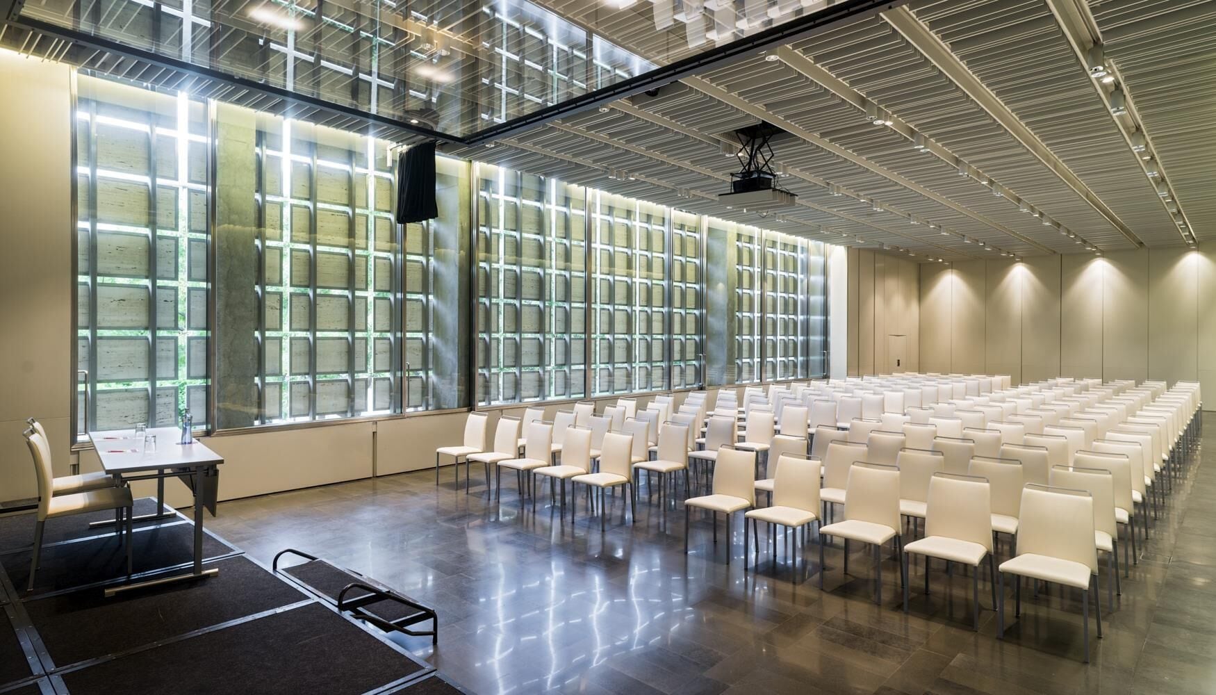 Un salón de conferencias con filas de sillas, una mesa con sillas y ventanas grandes con diseño de cuadrícula.