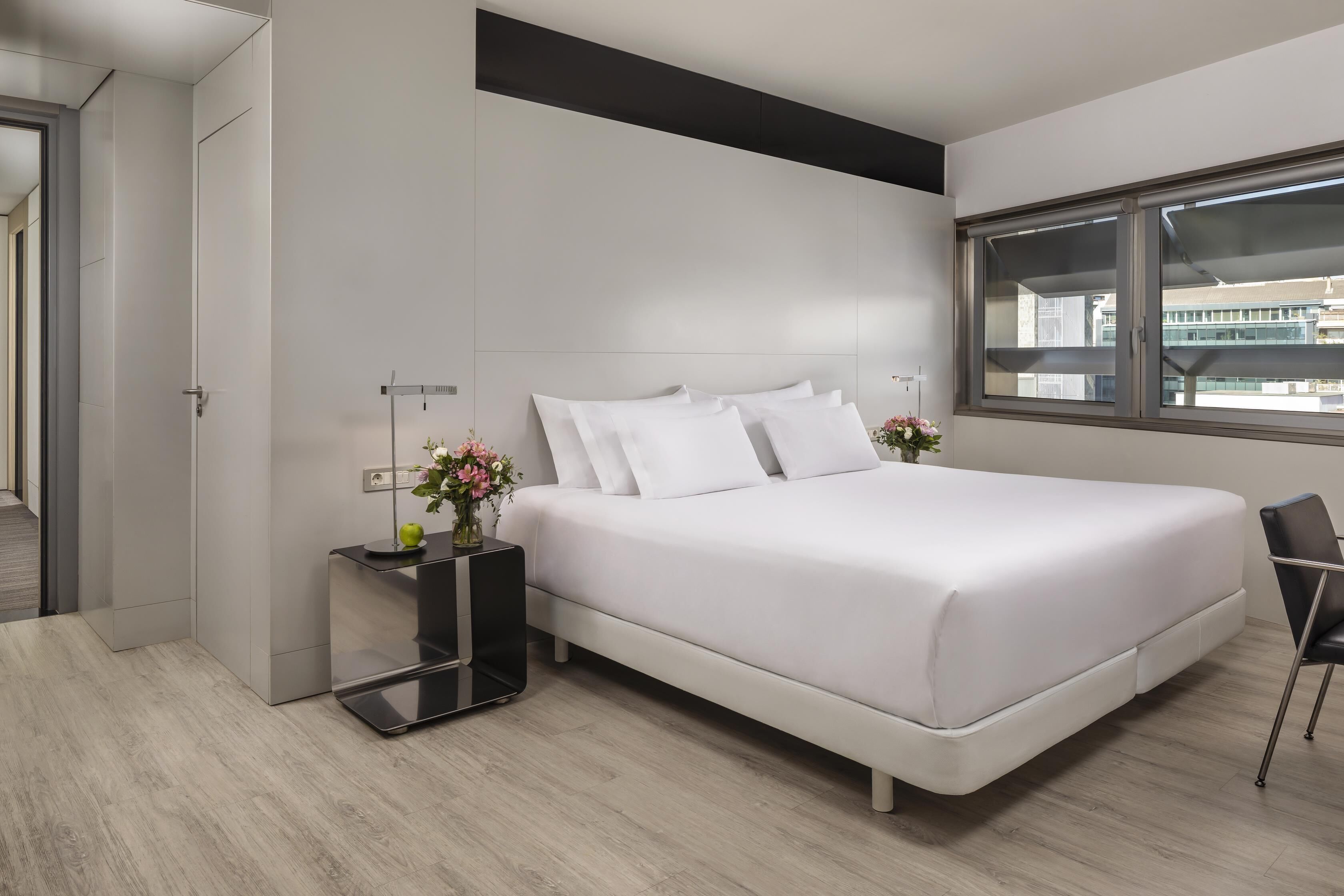 Un dormitorio moderno con una cama grande, una mesita de noche con un jarrón y una ventana con vista a la ciudad.