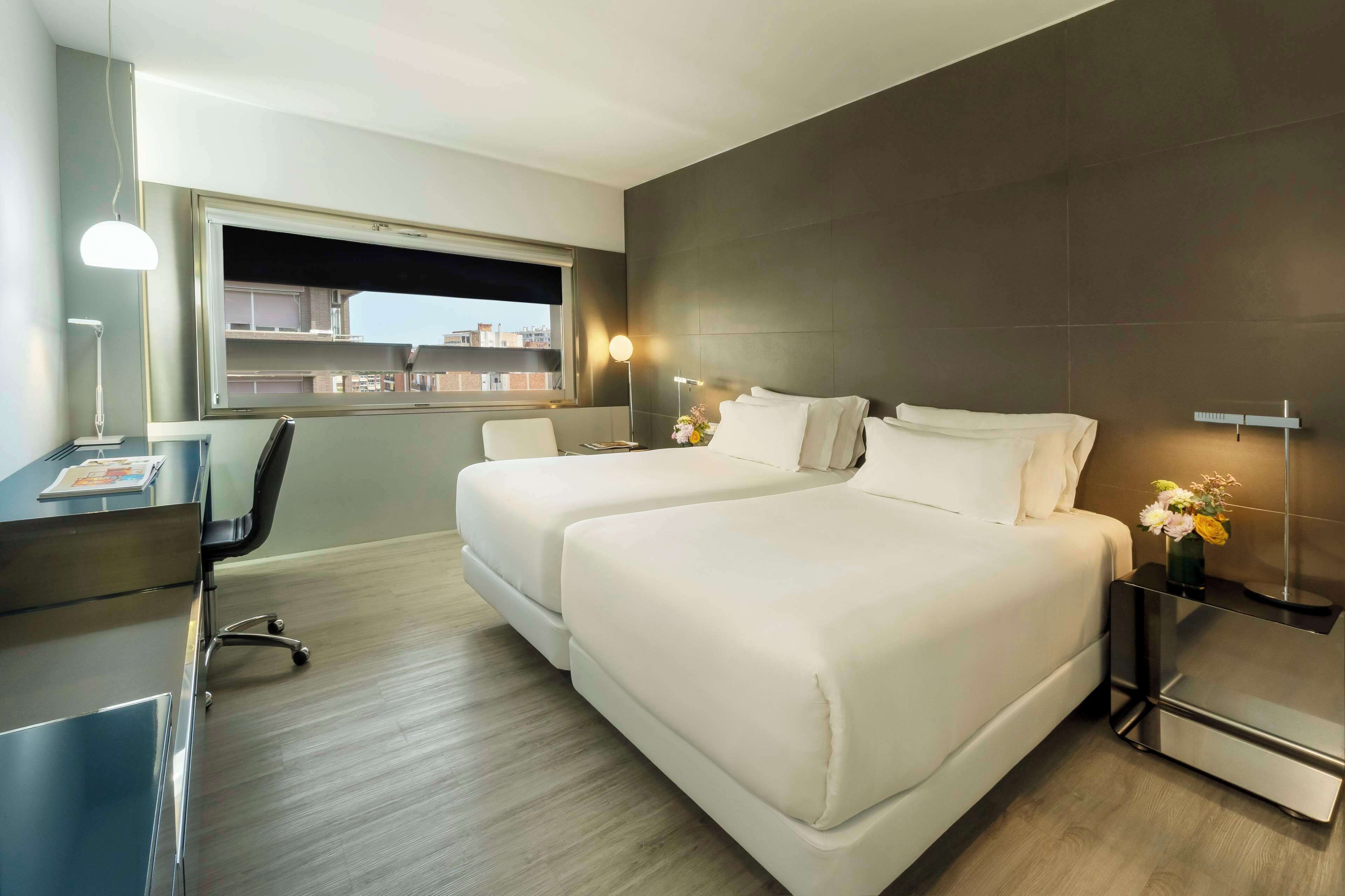 Habitación de hotel moderna con una cama grande, un escritorio y una silla. Se ve una ventana con vistas a edificios y un cielo nocturno.