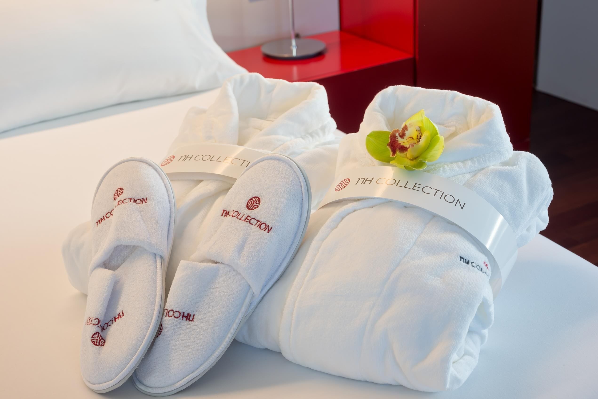 A neatly made bed with white bedding and a pair of white slippers with a logo.