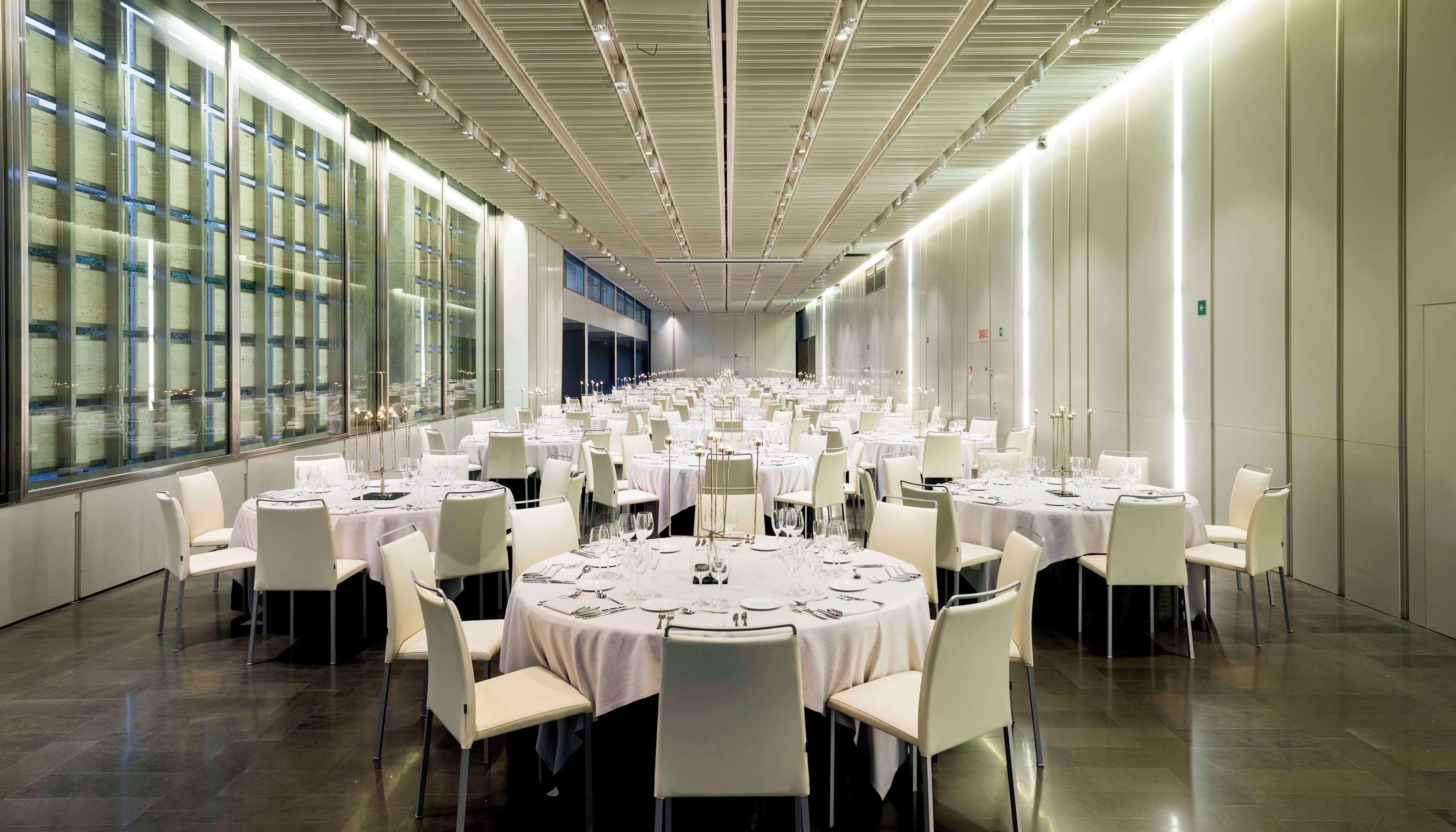 A large banquet hall with round tables and chairs, high ceilings with linear lighting, and large windows along one wall.