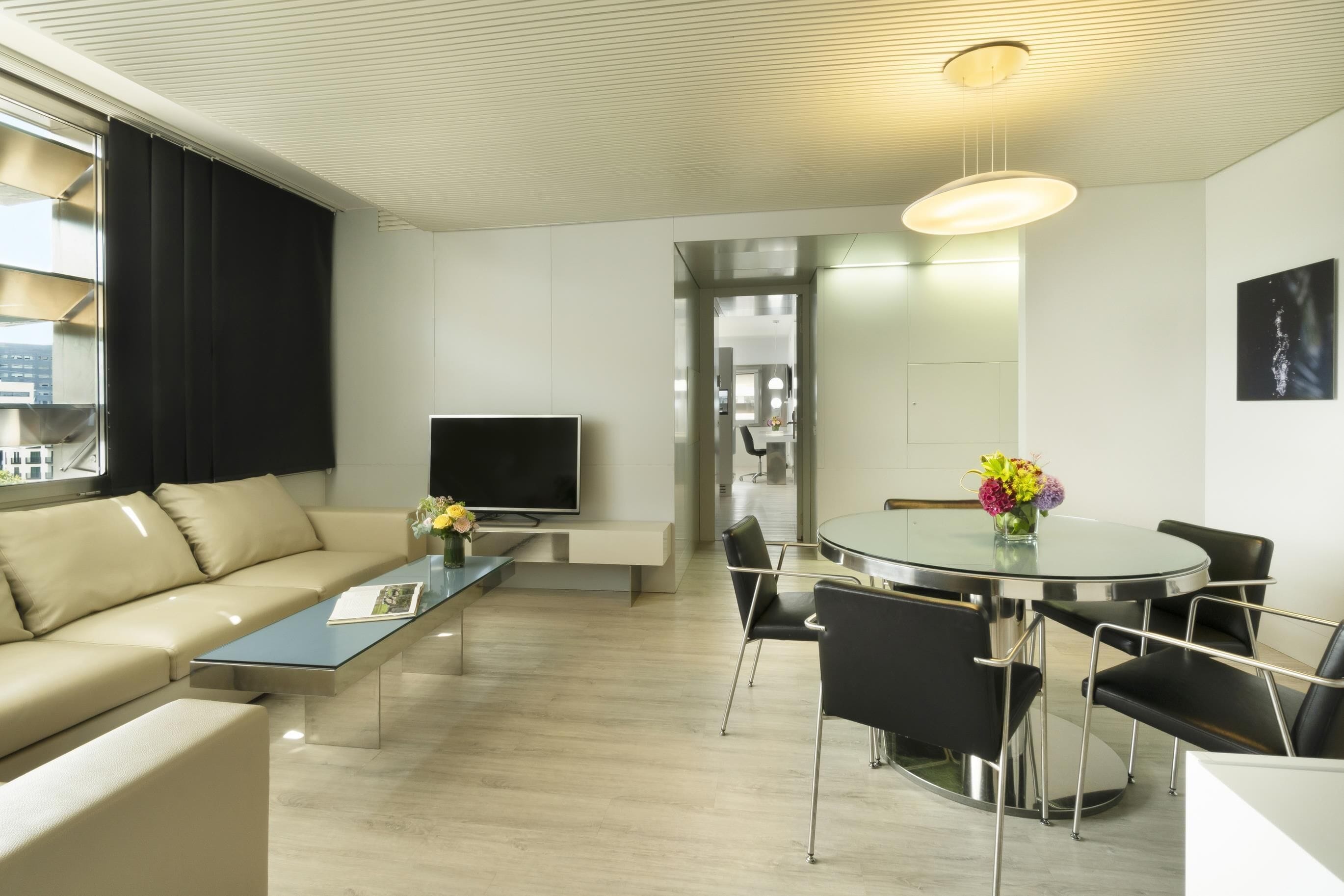 A modern living room with a sofa, coffee table, TV, dining table with chairs, and a large window with curtains.