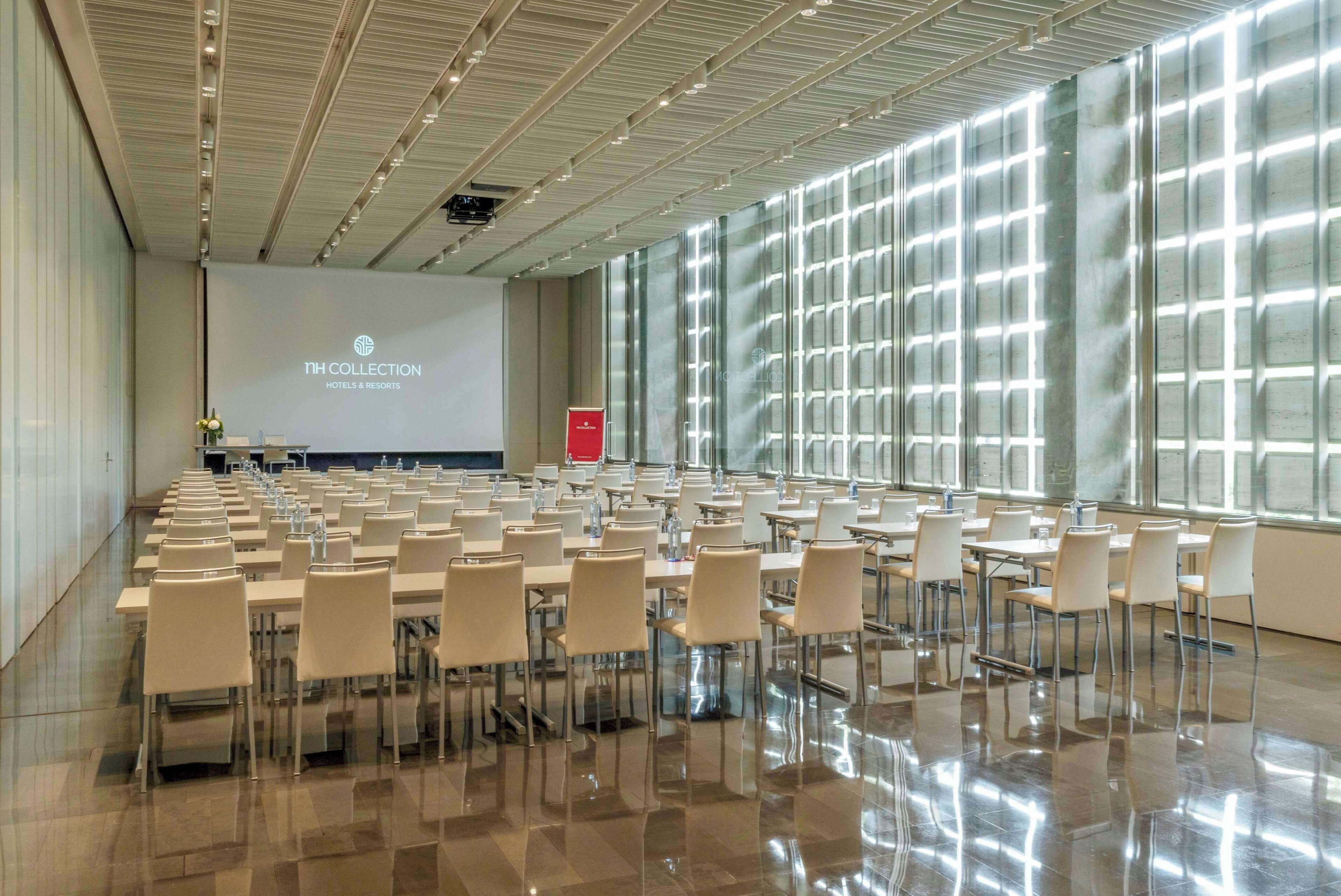Un amplio salón de conferencias con filas de mesas y sillas, una pantalla de proyección y un muro de cristal con diseño de cuadrícula.