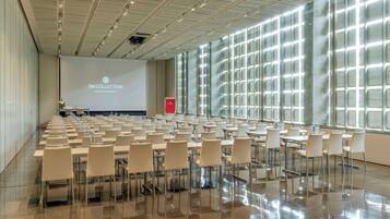 Un amplio salón de conferencias con filas de mesas y sillas, una pantalla de proyección y un muro de cristal con diseño de cuadrícula.