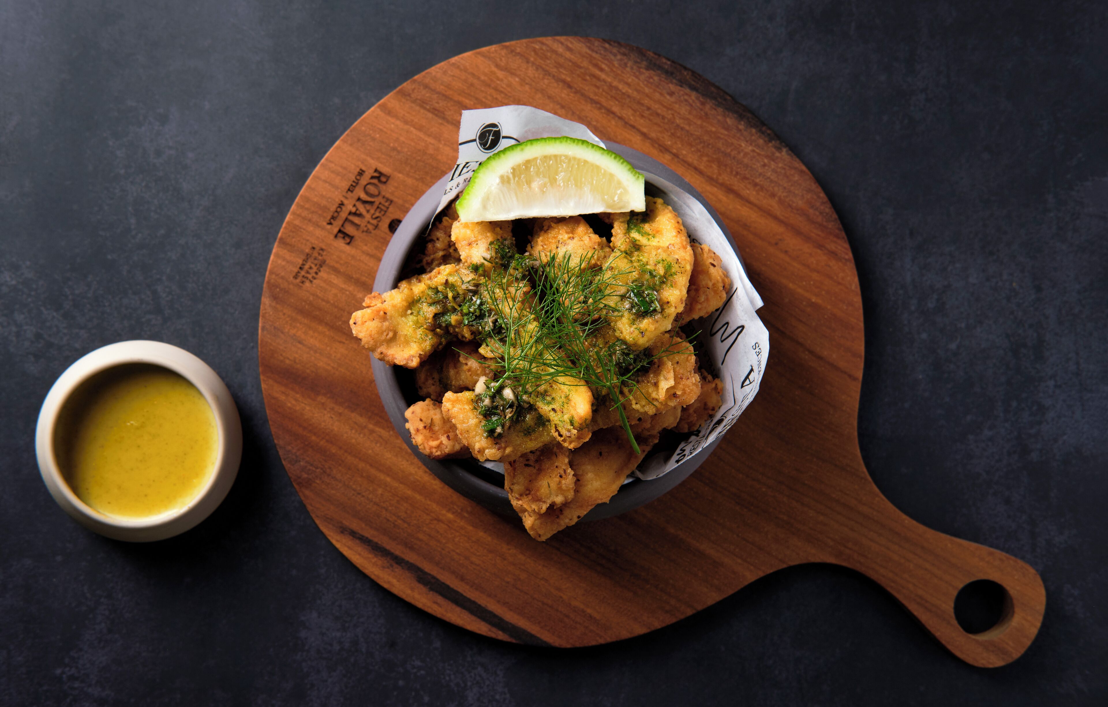 A wooden bowl with fried food garnished with herbs and a lime slice, accompanied by a small bowl of sauce on a dark surface.