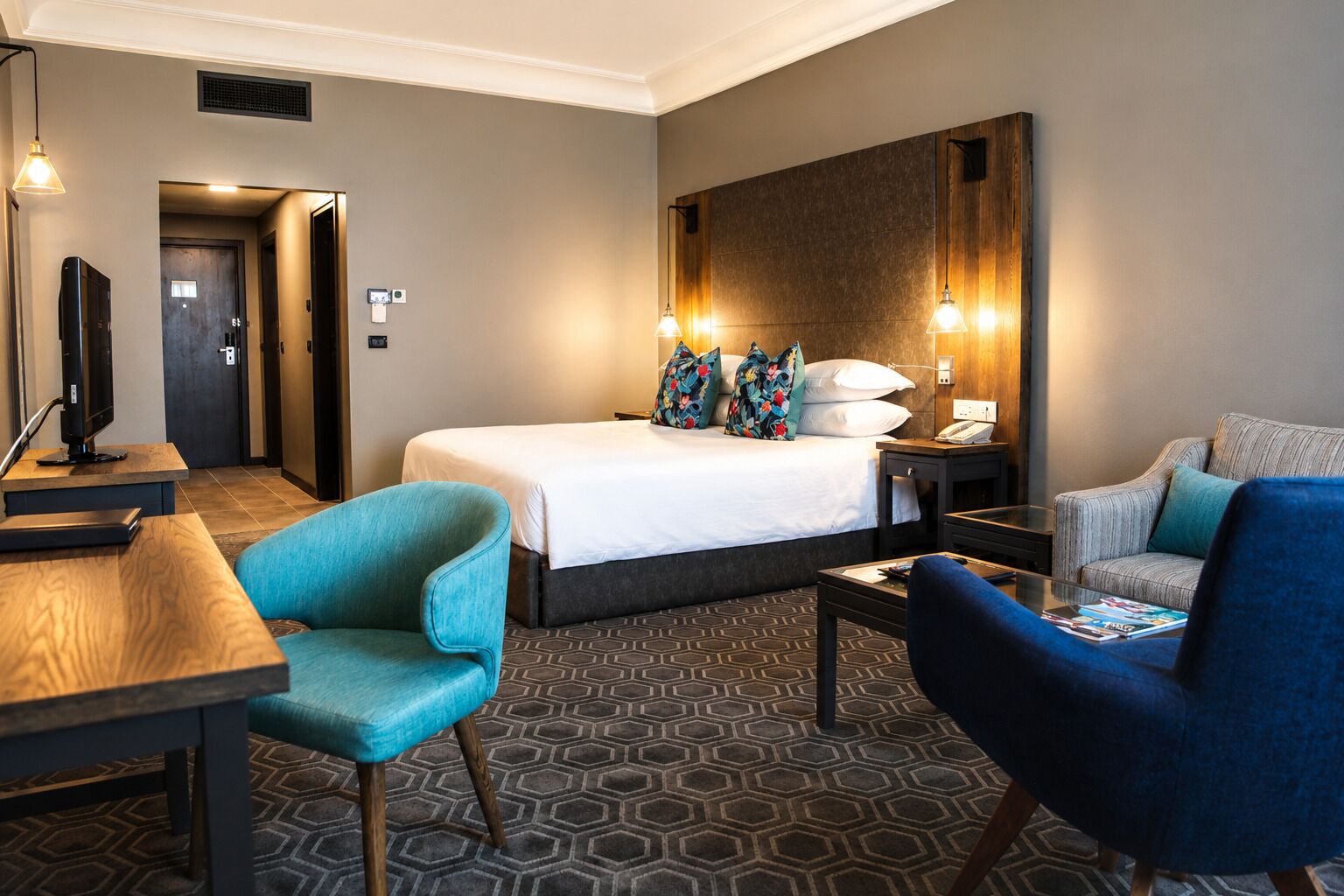 A hotel room with a bed, two chairs, a desk, and a television.