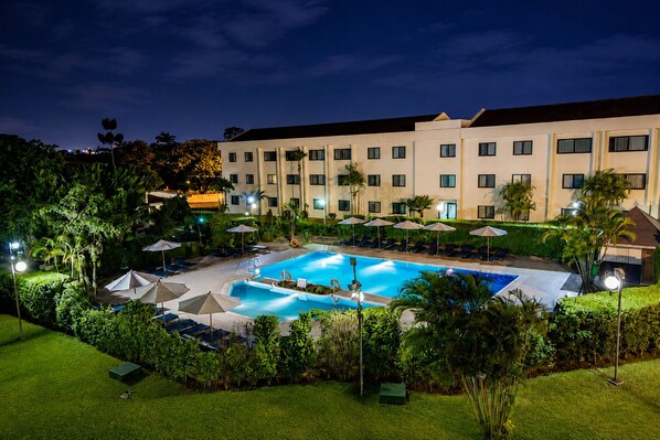 A hotel with a swimming pool surrounded by lounge chairs and umbrellas, set against a backdrop of a multi-story building. - Fiesta Royale Hotel (Accra)