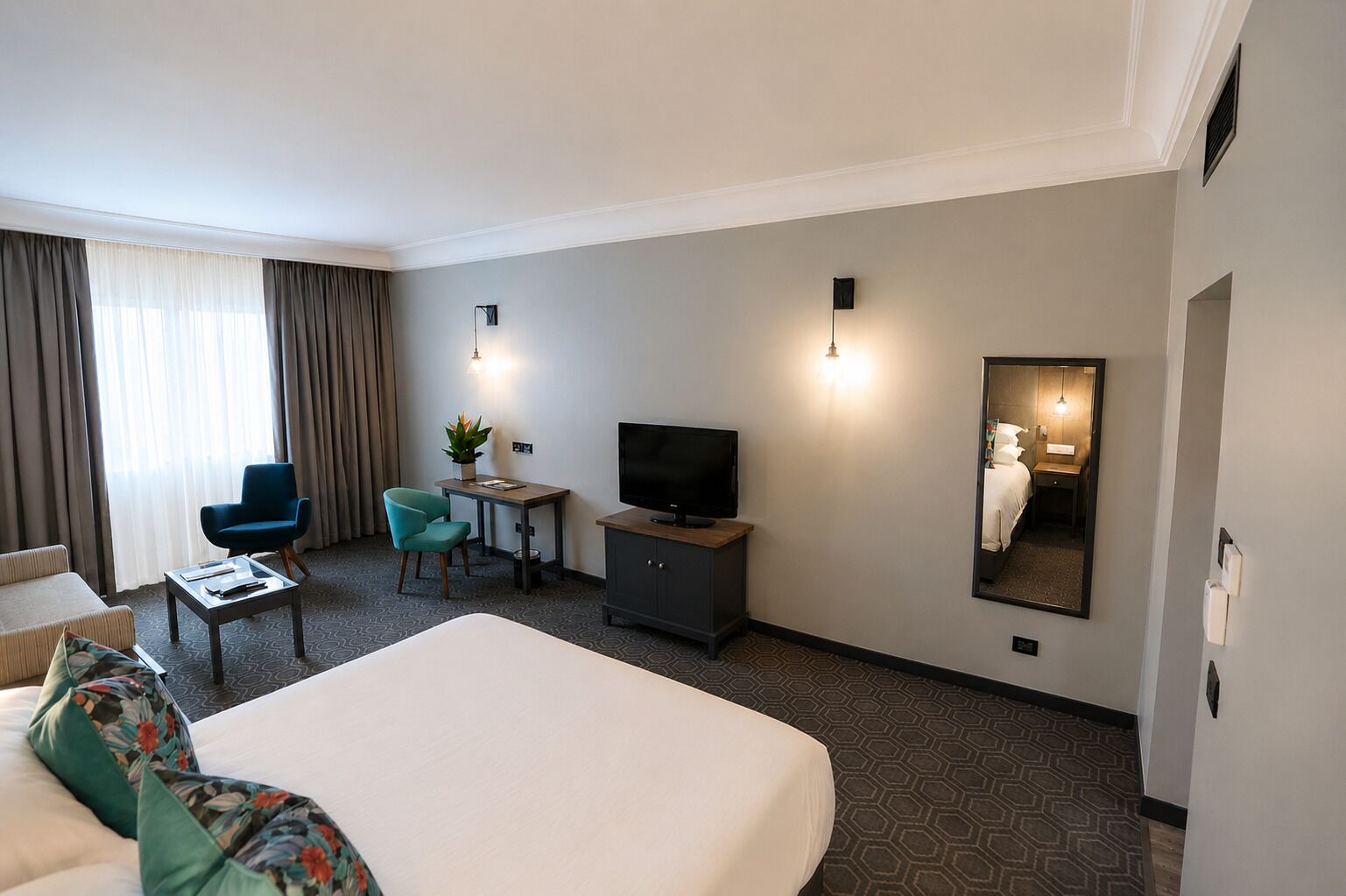 Une chambre d’hôtel avec un grand lit, un bureau avec une télévision, une chaise, une petite table avec une plante et un miroir au mur.