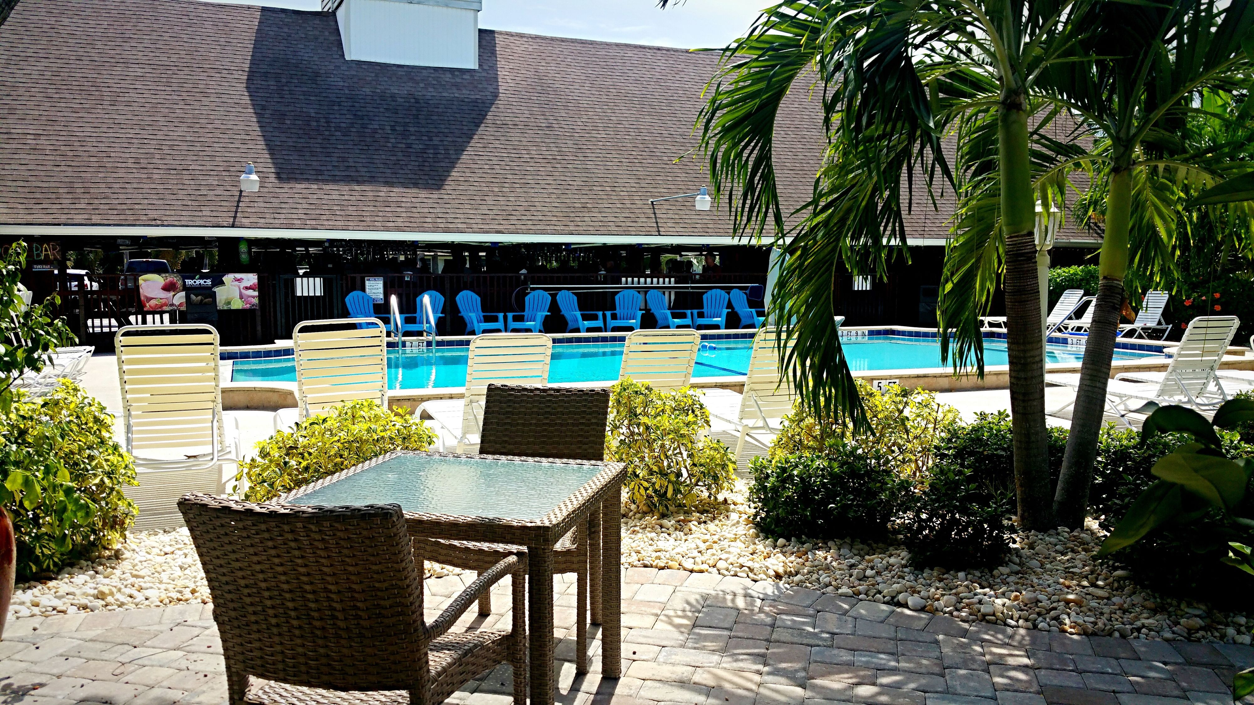 A pool area with lounge chairs and a table, surrounded by greenery and a building with a tiled roof.