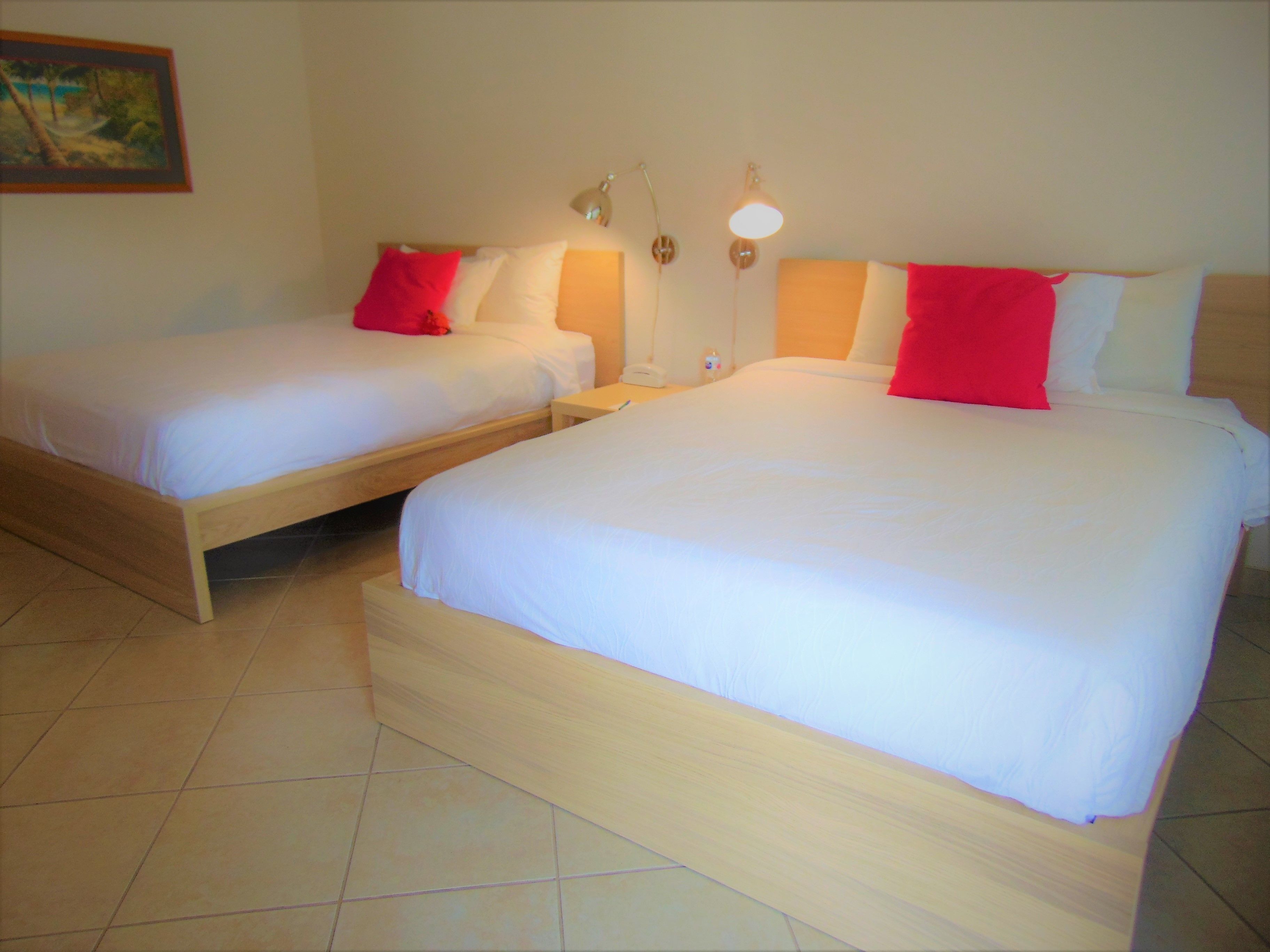 Two single beds with wooden frames and white bedding, each with a red pillow and a bedside table with a lamp.