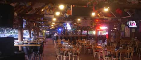 Un restaurant intérieur avec des tables et des chaises en bois, un coin bar et diverses décorations, notamment des drapeaux et des guirlandes lumineuses.