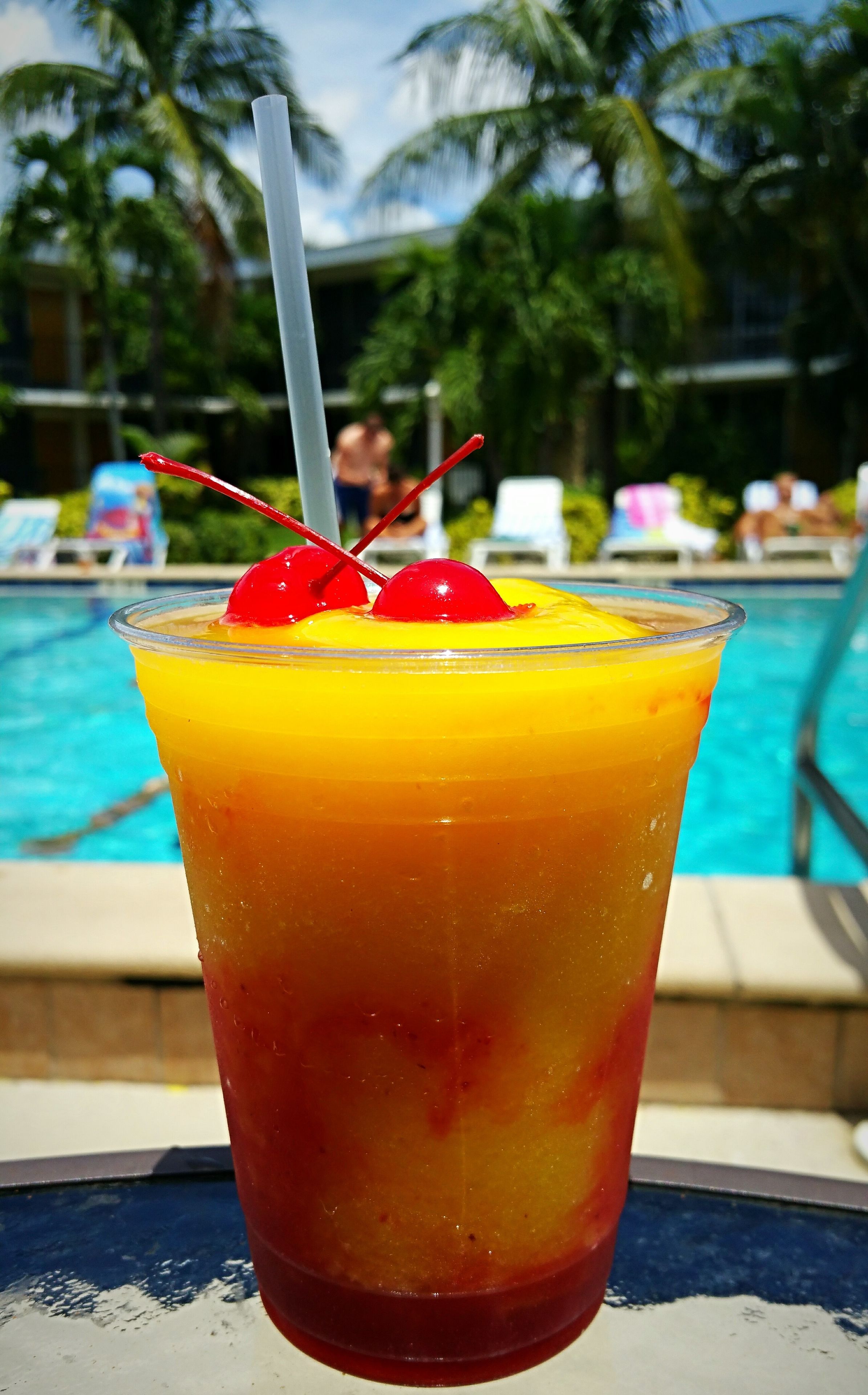 Un cocktail tropical servi avec une paille et une cerise sur le bord du verre, posé devant une piscine, avec des chaises longues et des palmiers en arrière-plan.