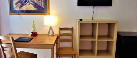 A dining area with a wooden table, two chairs, a painting, a TV mounted on the wall, and a small cabinet.
