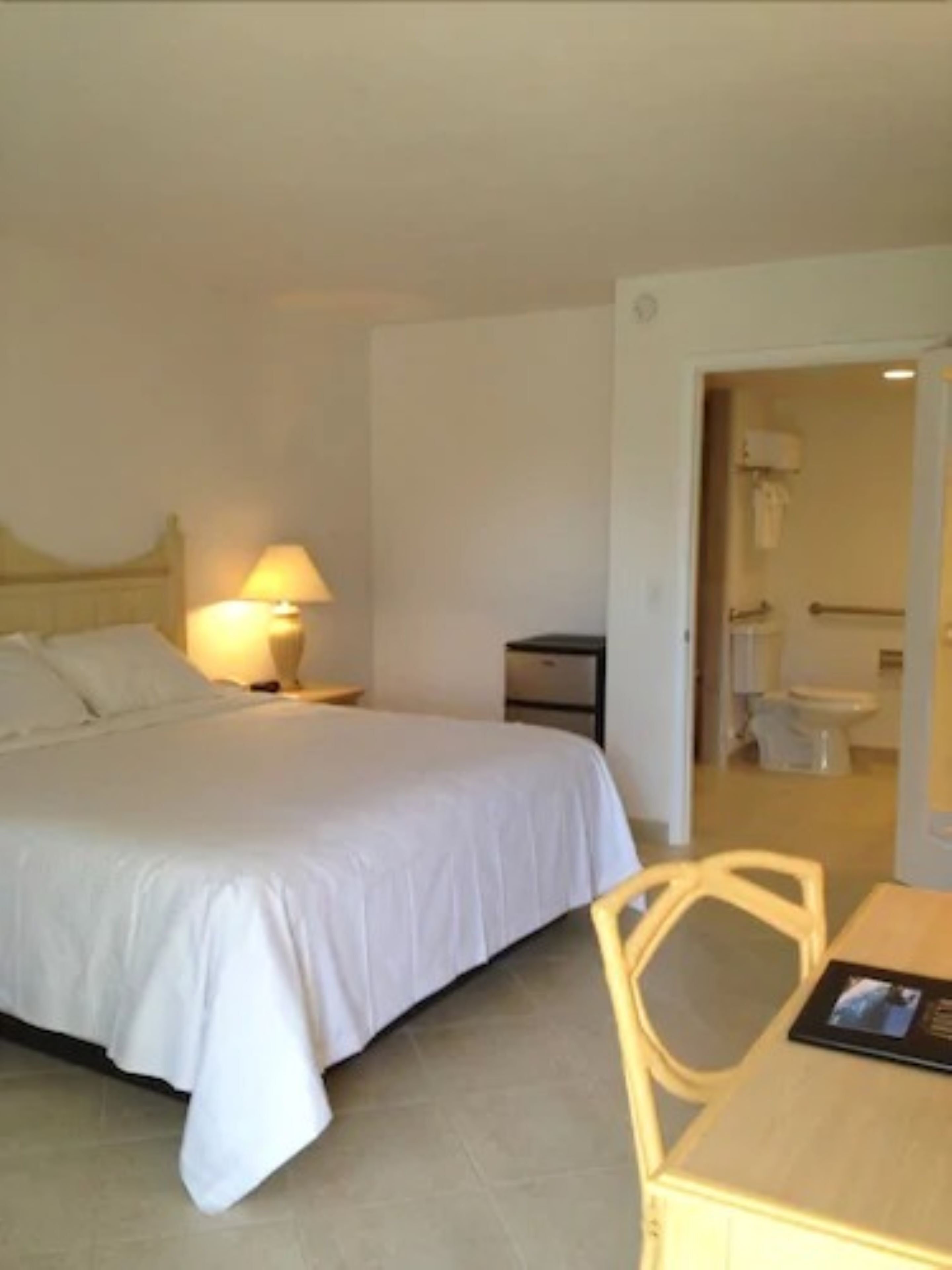 Een hotelkamer met een bed, een nachtkastje met een lamp, een stoel en een klein tafeltje met onderzetter.