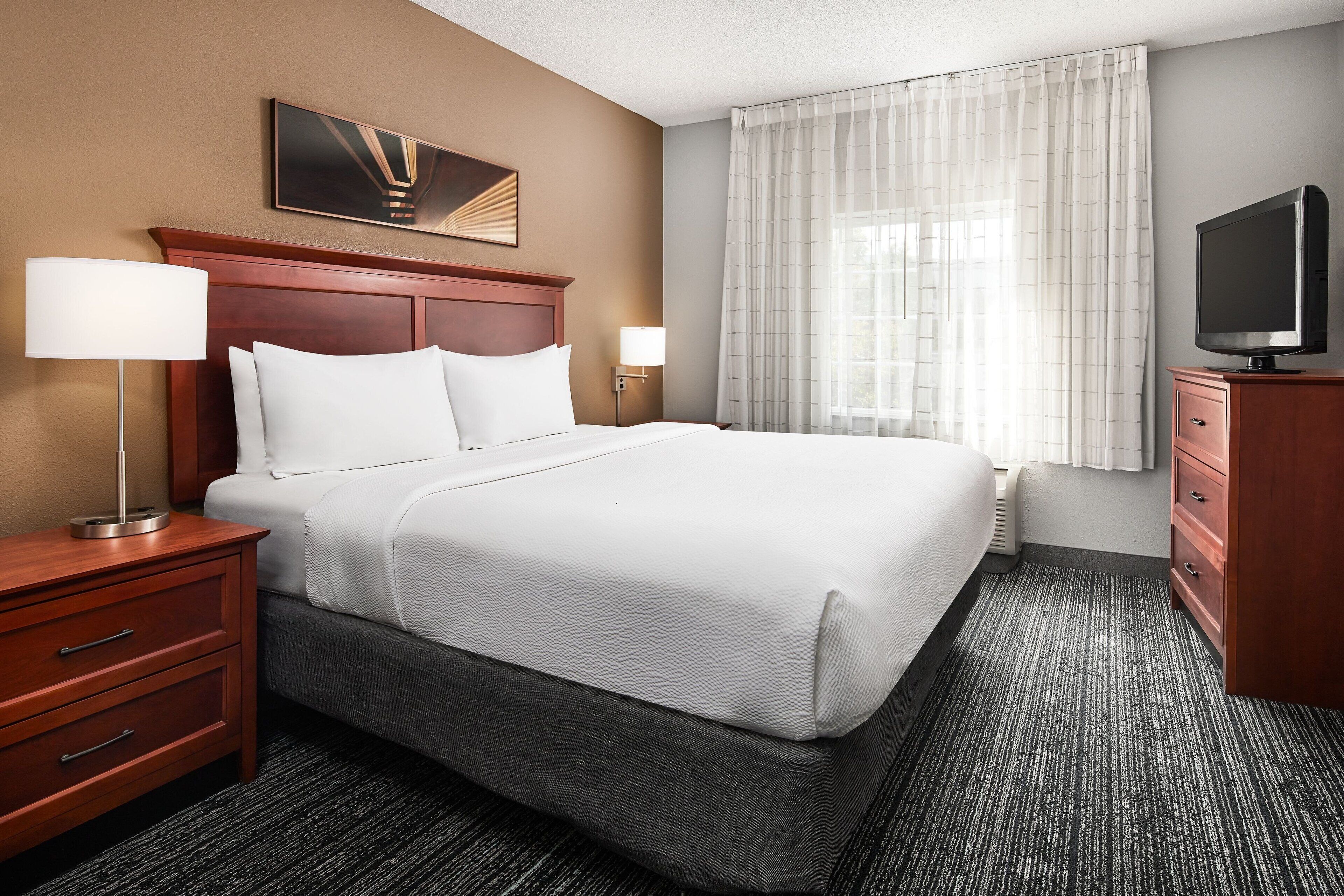 A hotel room with a large bed, wooden headboard, two bedside tables with lamps, a flat-screen TV, and a window with sheer curtains.