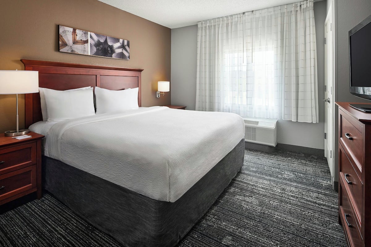 A hotel room with a large bed, a wooden headboard, two bedside tables with lamps, a flat-screen TV, and a window with curtains.
