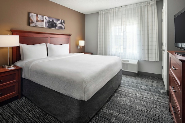 A hotel room with a large bed, a wooden headboard, two bedside tables with lamps, a flat-screen TV, and a window with curtains.