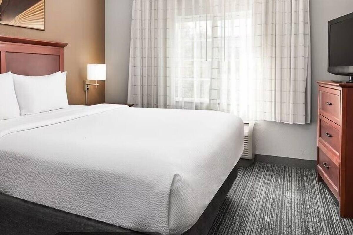 A hotel room with a large bed, a wooden headboard, a nightstand with a lamp, a dresser, and a television on a stand.