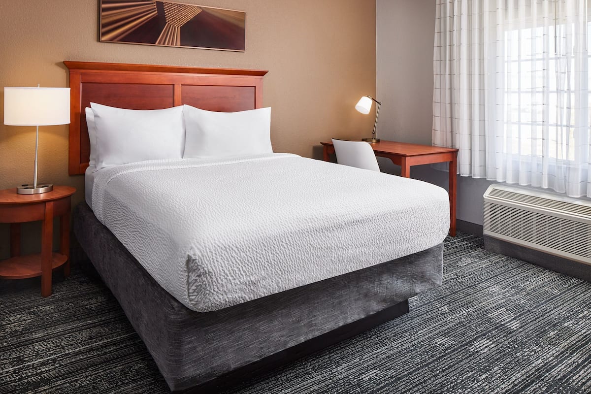 A hotel room with a large bed, two bedside tables, a lamp, a heater, and a window with curtains.