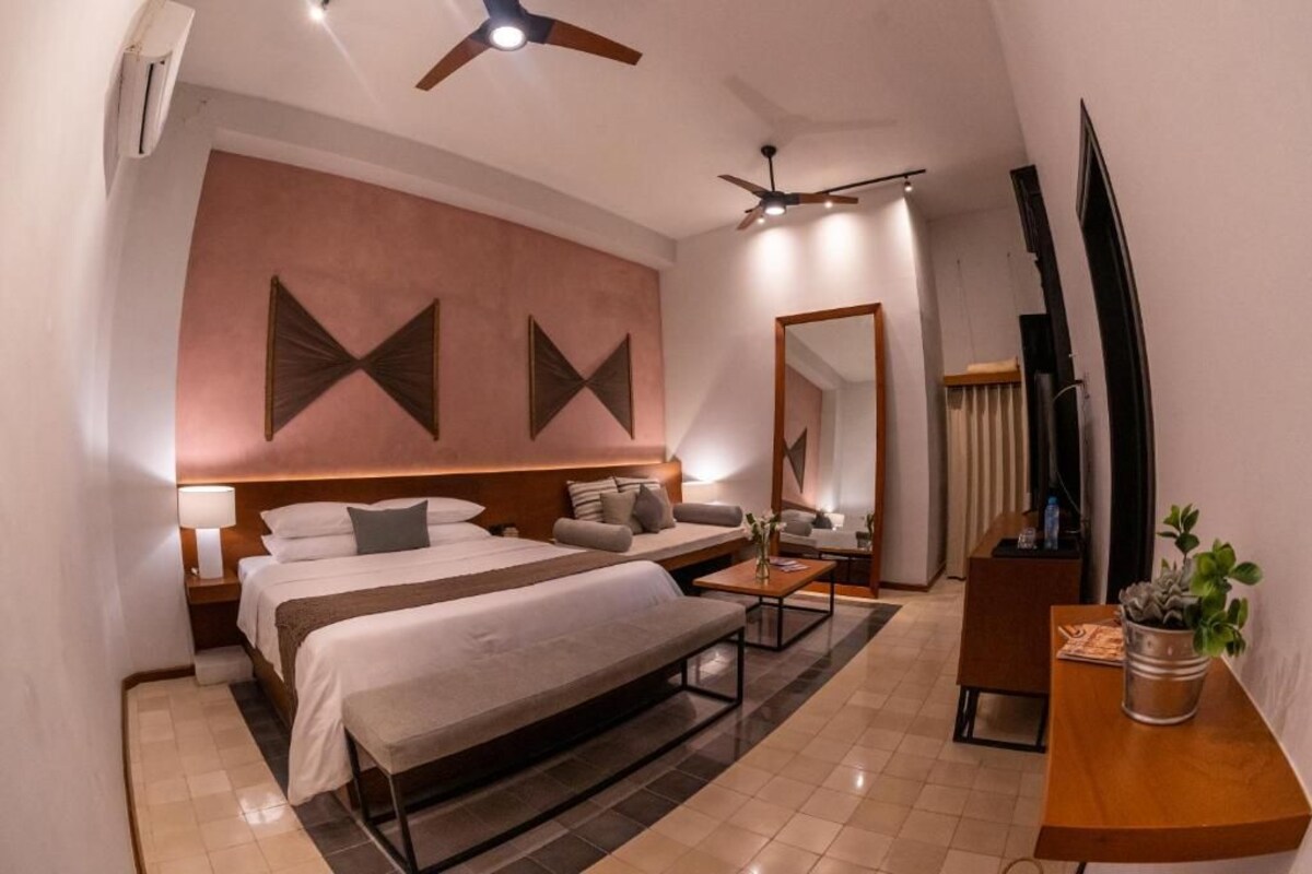 Habitación de hotel con cama, sofá, escritorio, espejo, televisor y ventilador de techo.
