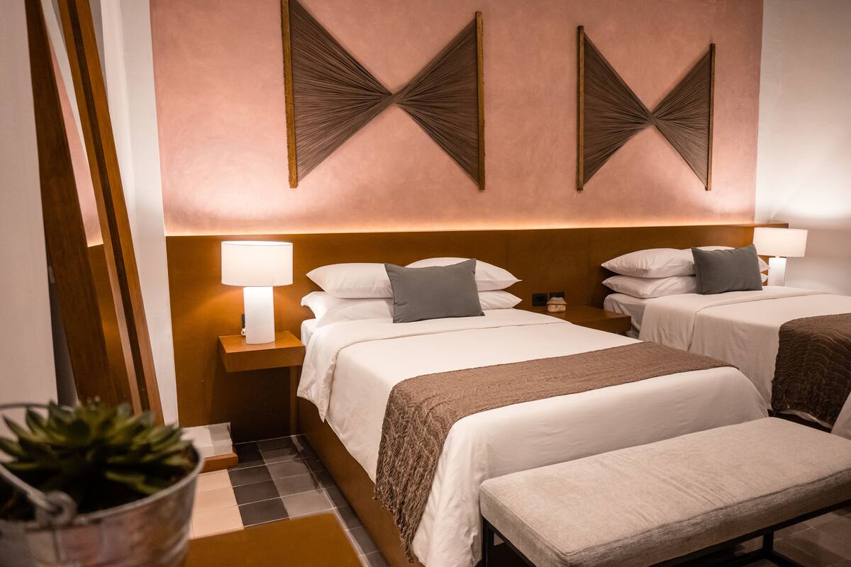 A hotel room with two beds, a bedside table with a lamp, a wall-mounted light fixture, and a potted plant.