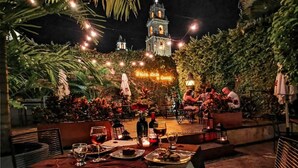 An outdoor dining area with tables set for a meal, surrounded by lush greenery and a view of a historic building with a clock tower.