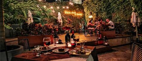 Un espace de restauration en plein air avec des tables dressées pour un repas, entouré d’une végétation luxuriante et offrant une vue sur un bâtiment historique doté d’un beffroi.
