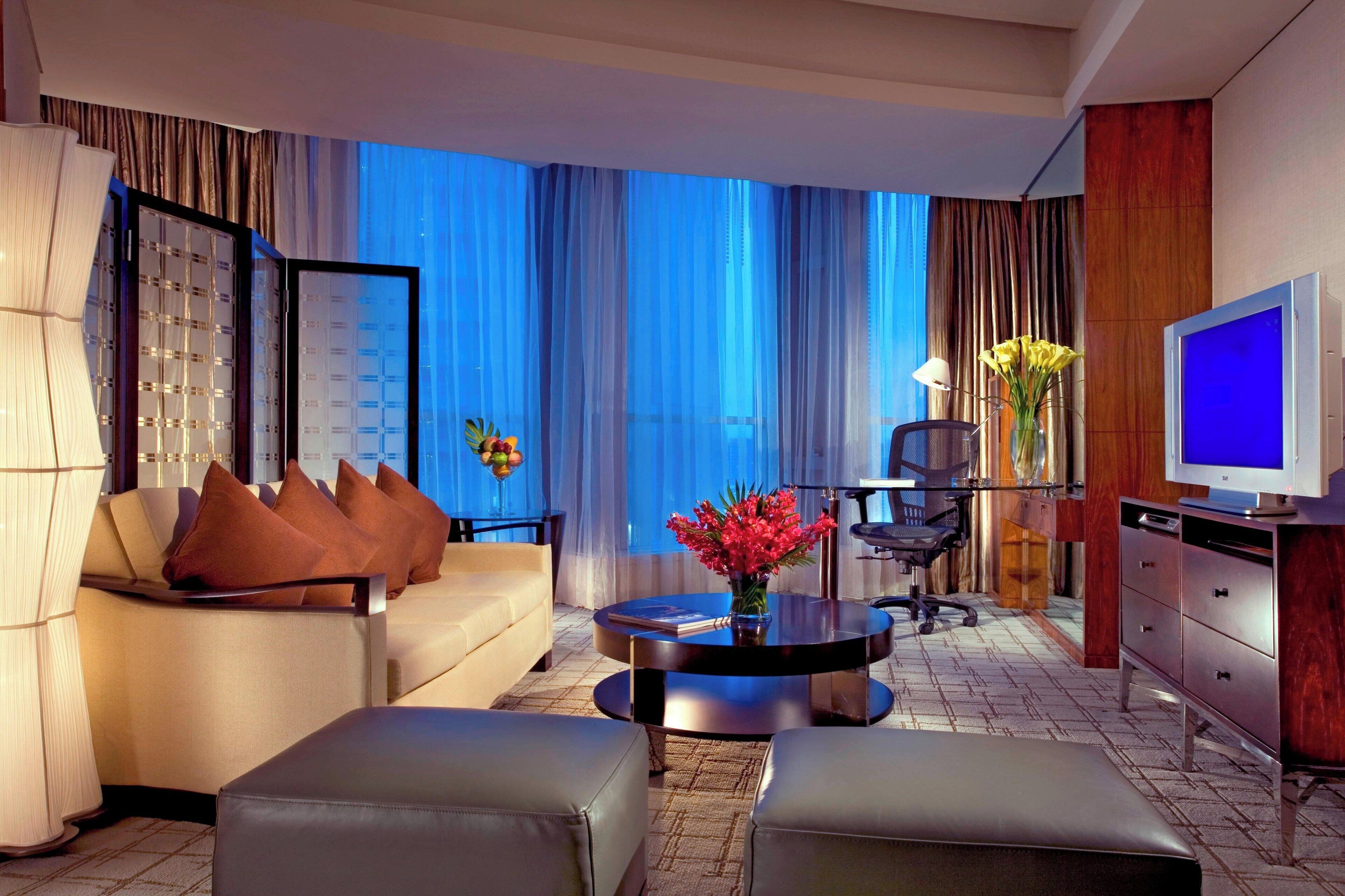 A modern hotel room with a sofa, ottoman, coffee table, TV, and a desk with a chair.