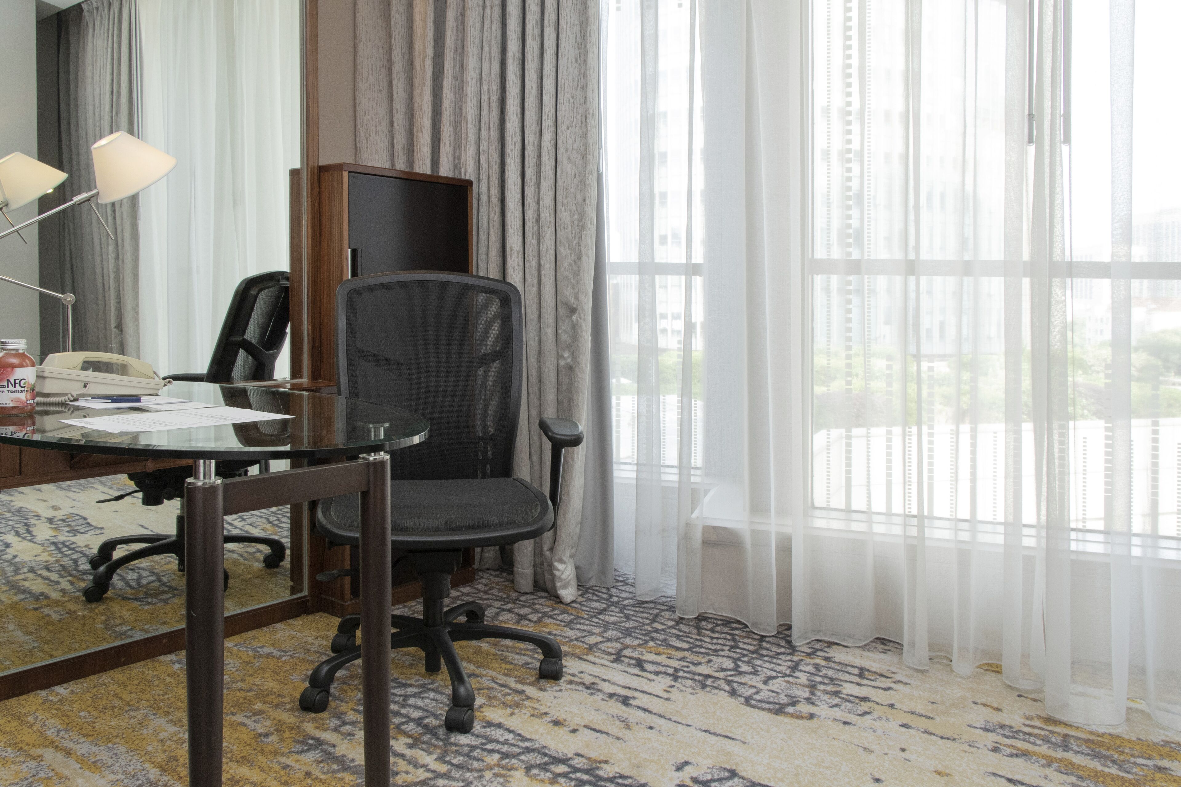A hotel room with a desk, office chair, lamp, and a large window with sheer curtains.